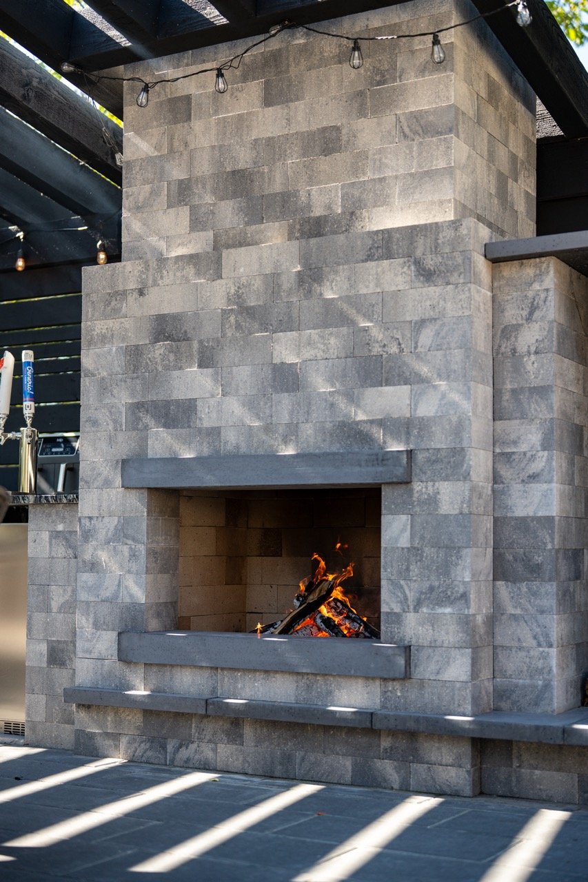 A stone fireplace with a fire burning inside, located outdoors with a shaded patio area.