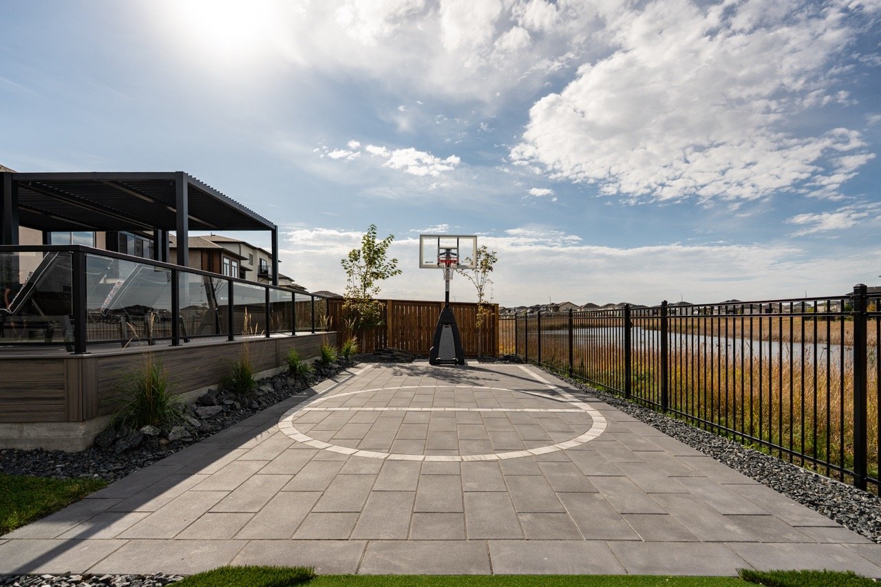 Backyard with a basketball hoop on a paved court, fencing on both sides, neighboring houses on the left, and a body of water beyond the fence on the right