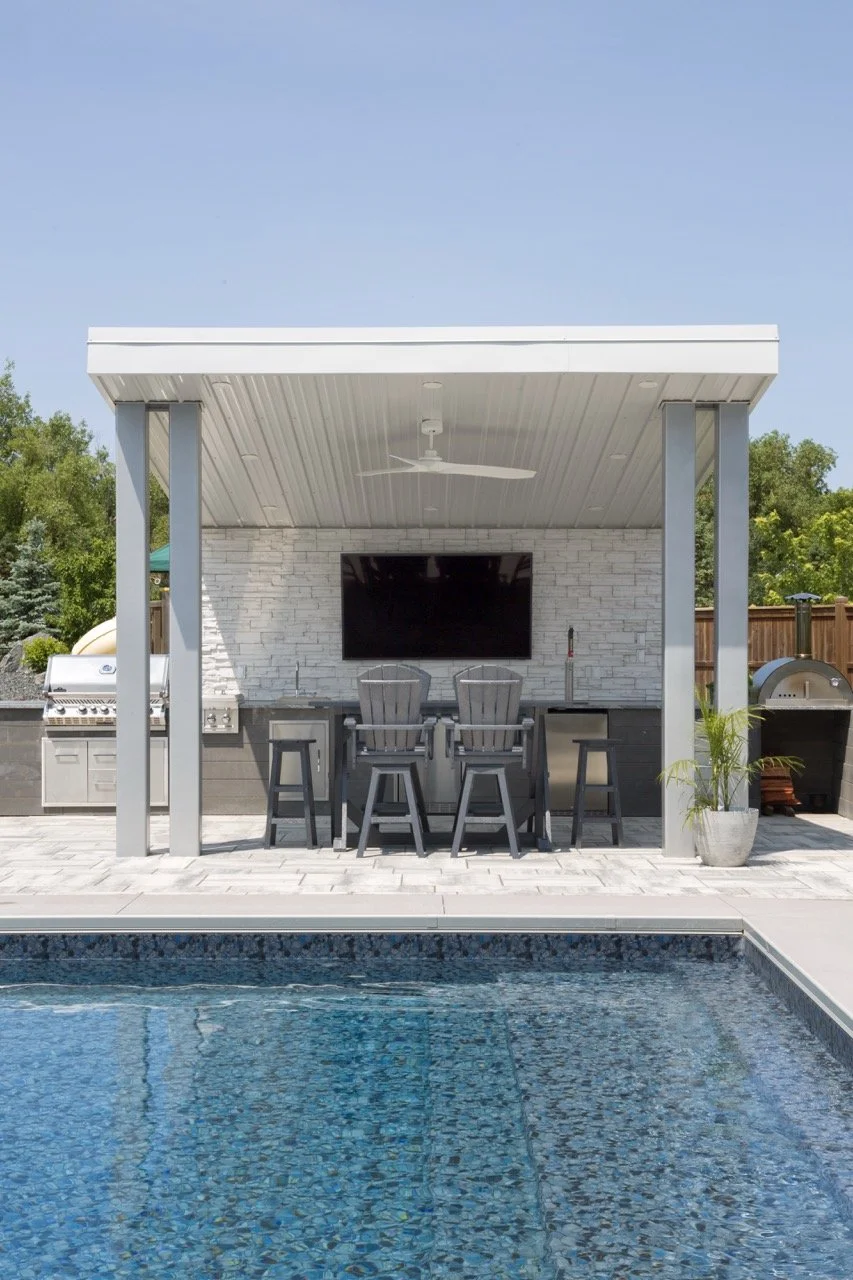 Outdoor kitchen and bar area with a pool in the foreground, featuring a countertop, bar stools, a brick wall, TV, and ceiling fan under a roofed structure.