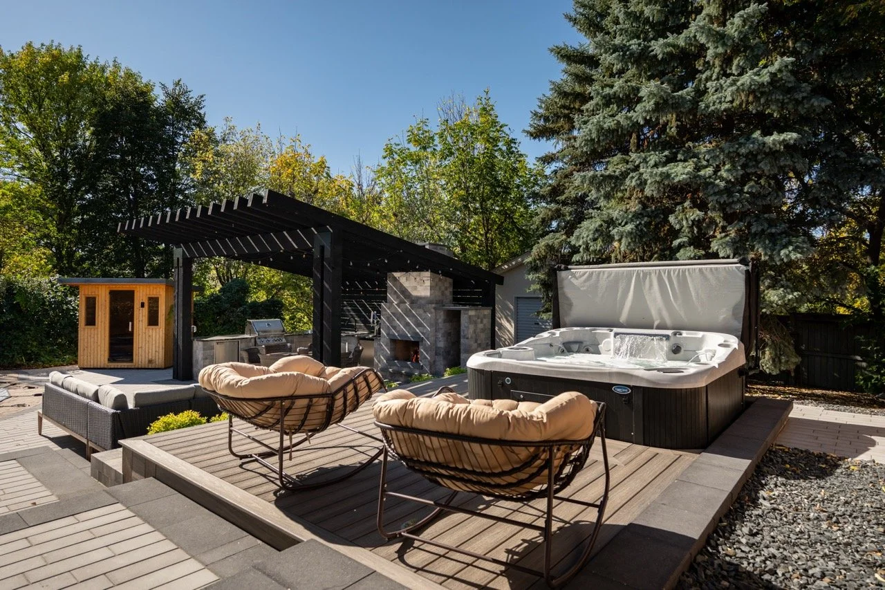Outdoor patio area with a hot tub, two cushioned lounge chairs, a seating area, a small wooden sauna, and a black pergola with a fireplace, surrounded by trees under a clear blue sky.