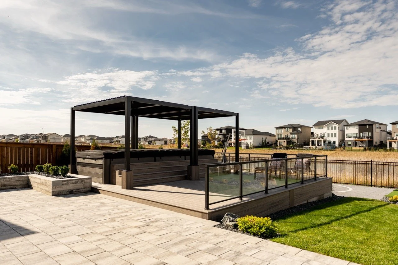 Backyard with a built-in hot tub under a black metal pergola, enclosed with glass fencing, surrounding a paved patio area, with houses in the distance and a partly cloudy sky.