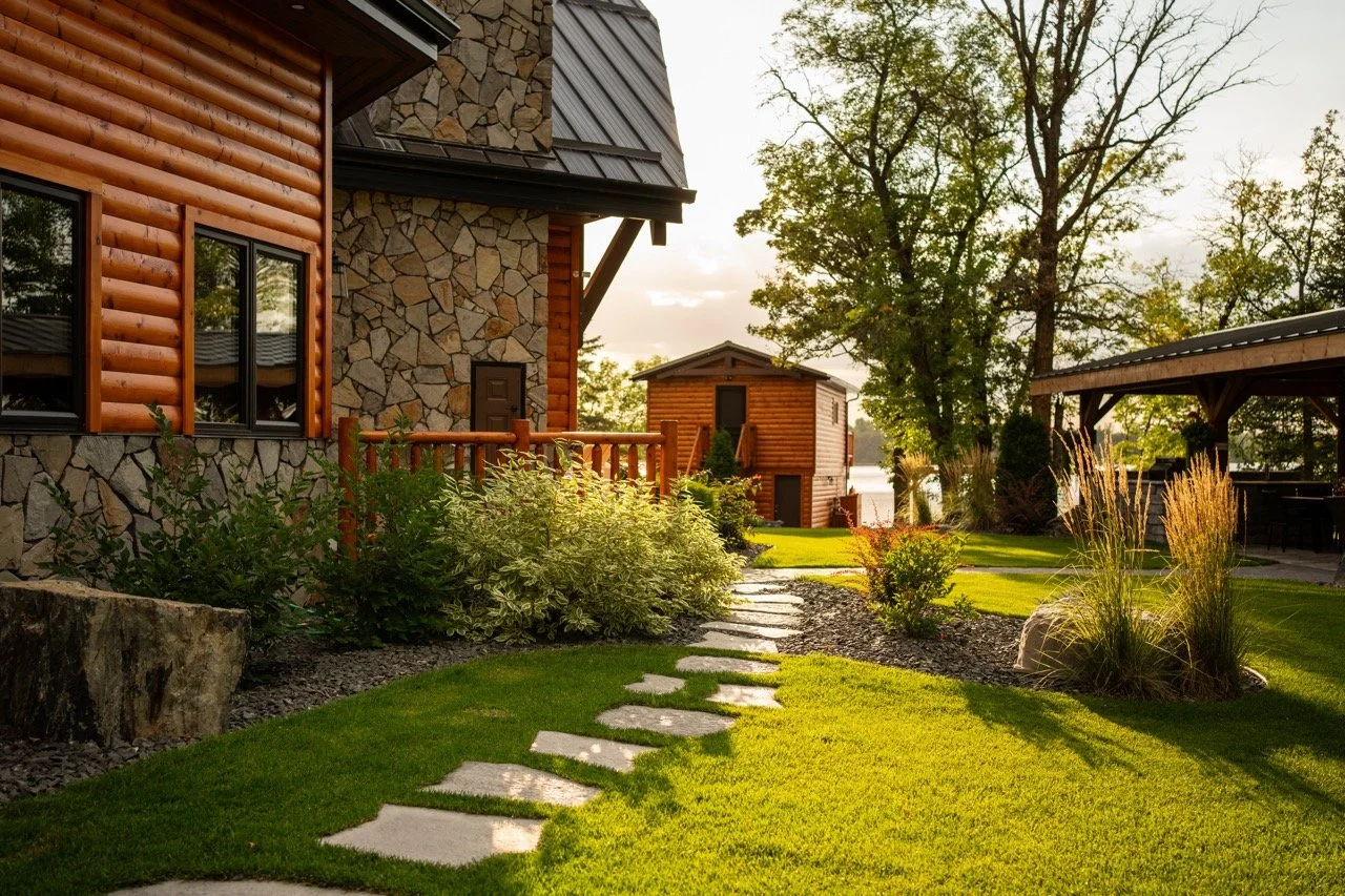 A well-maintained backyard garden with a green lawn, a stone pathway, various shrubs, and trees, with a wooden and stone house in the background during sunset.