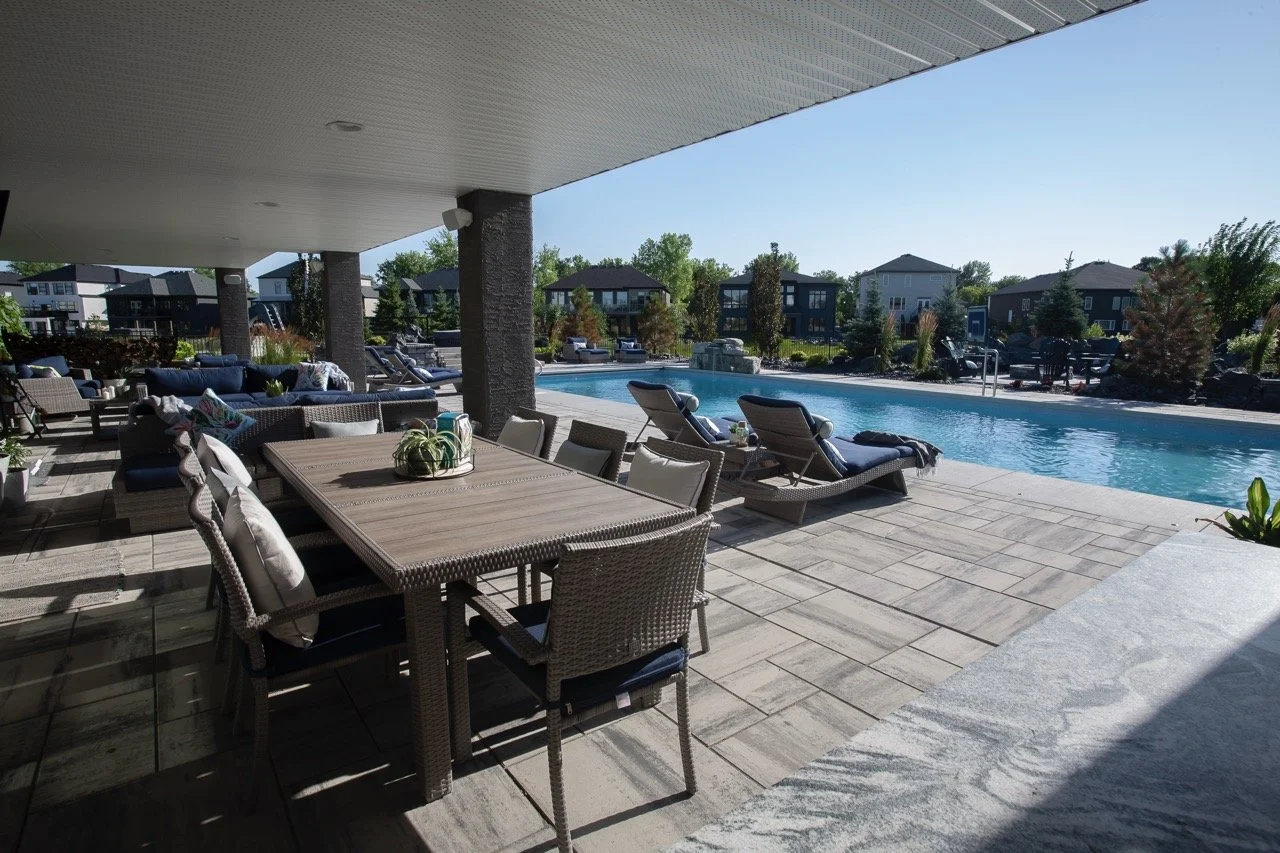Outdoor patio with dining table and chairs, lounge chairs by a swimming pool, surrounded by greenery and residential buildings.
