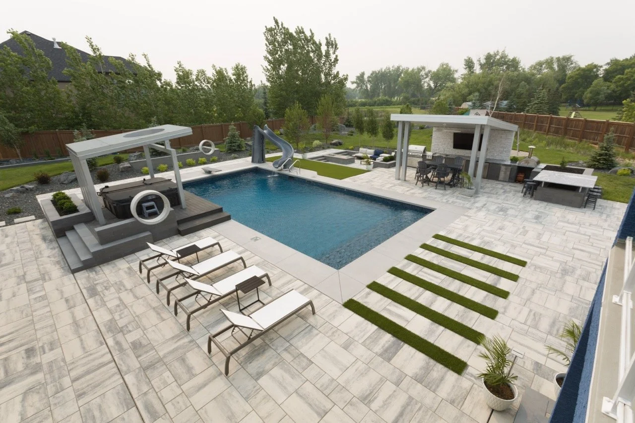 Luxurious backyard with swimming pool, lounge chairs, water slide, pergola seating area, and outdoor kitchen on a stone patio surrounded by greenery.