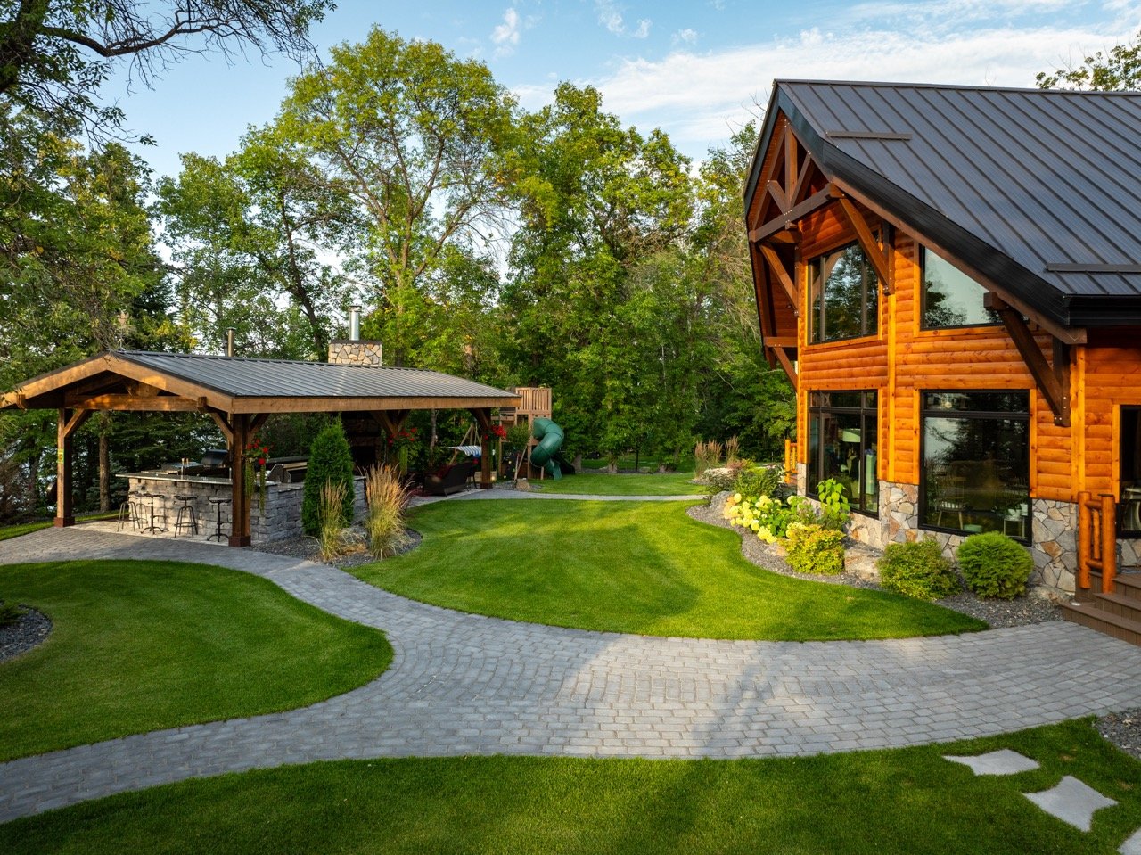 A backyard with a paved stone path, green lawn, and a large wooden log cabin-style house with large windows. There is also a covered outdoor seating area with a stone counter and bar stools, surrounded by trees and plants.