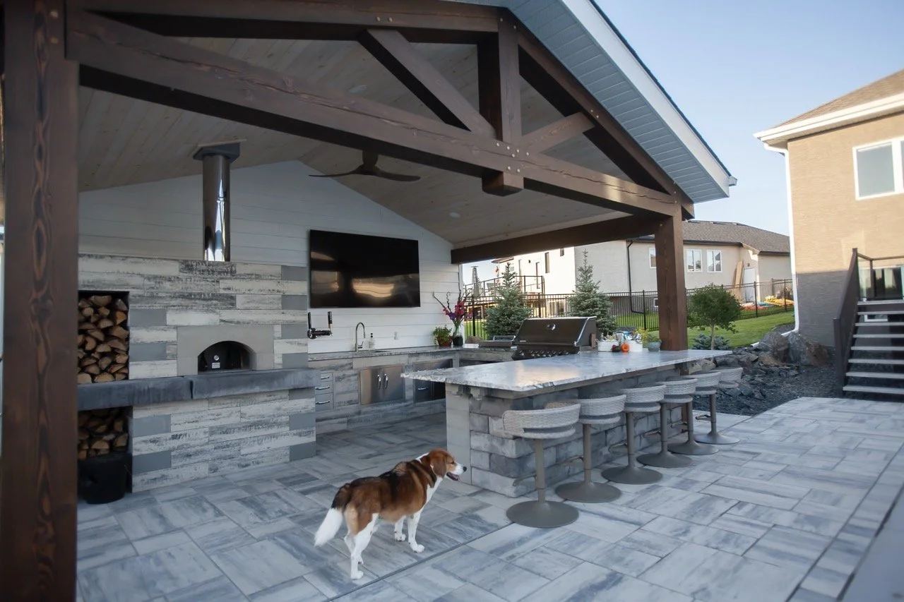 Outdoor kitchen with bar stools, a grill, pizza oven, stacked firewood, and a dog on a tiled patio.