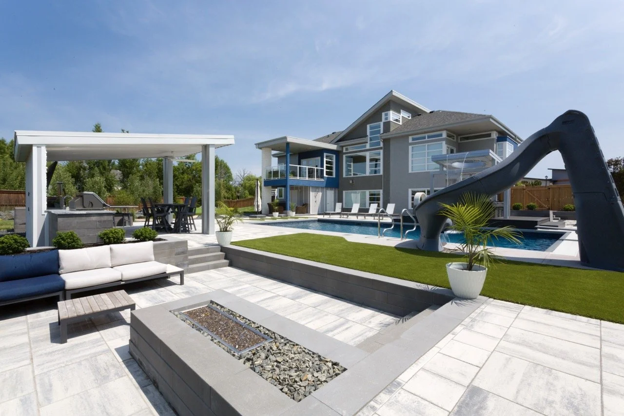 Modern backyard with pool, slide, patio seating, pergola, and outdoor kitchen, adjacent to a contemporary house on a sunny day.
