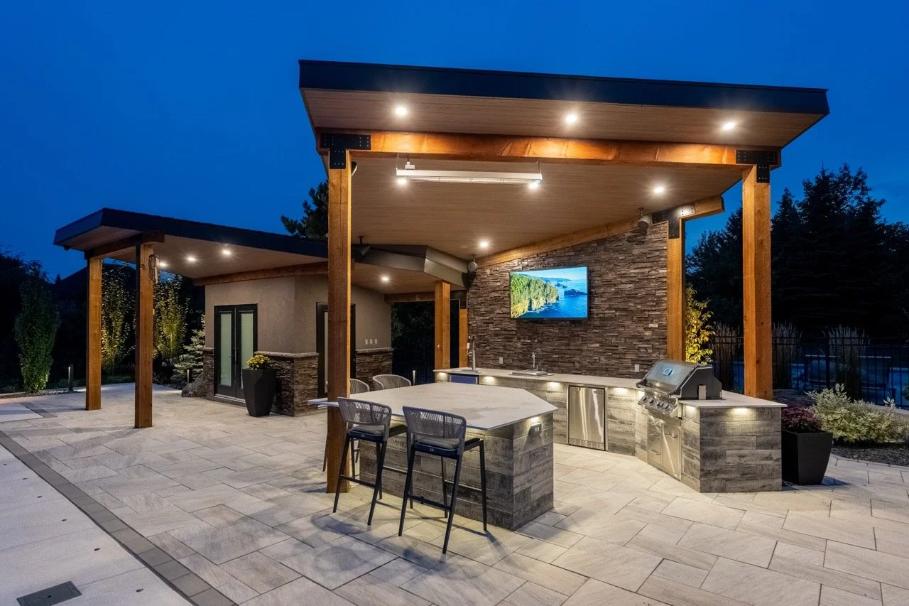 Outdoor kitchen with modern design, featuring a grill, sink, bar seating, and stone accents, illuminated by evening lighting.