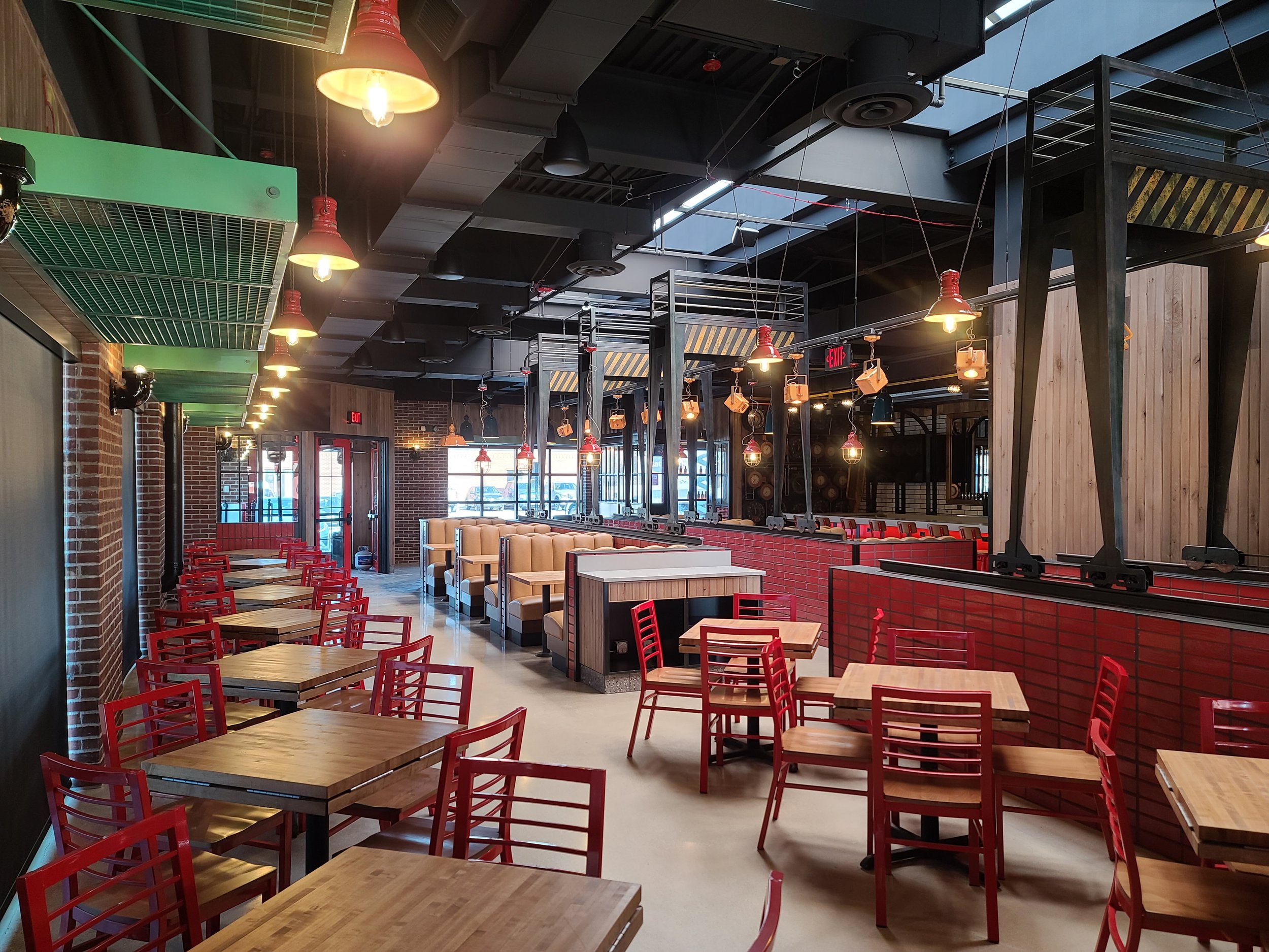 Interior of a modern restaurant with red chairs, wooden tables, exposed brick walls, and industrial-style pendant lighting.