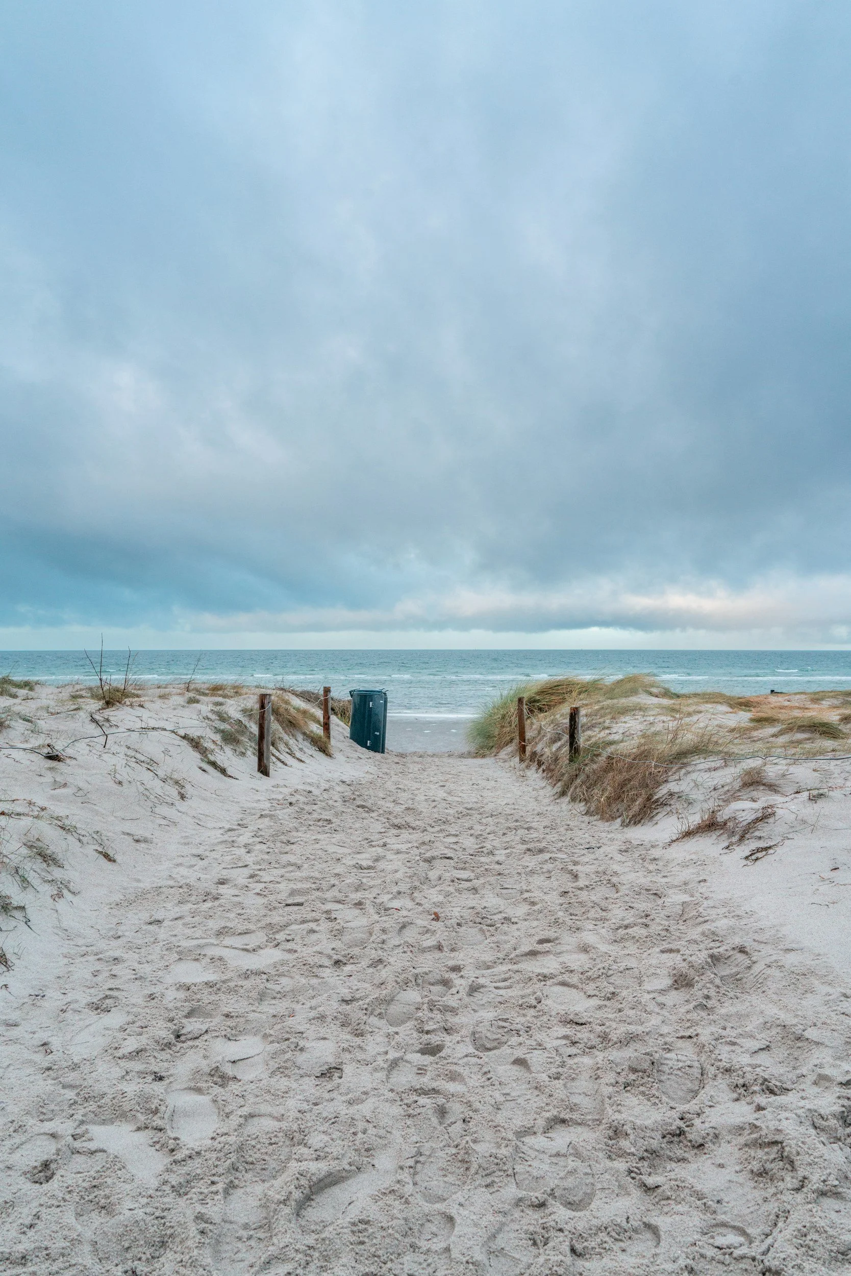 Sand pathway through dunes leading to the beach with an ocean view, cloudy sky, and a trash can.