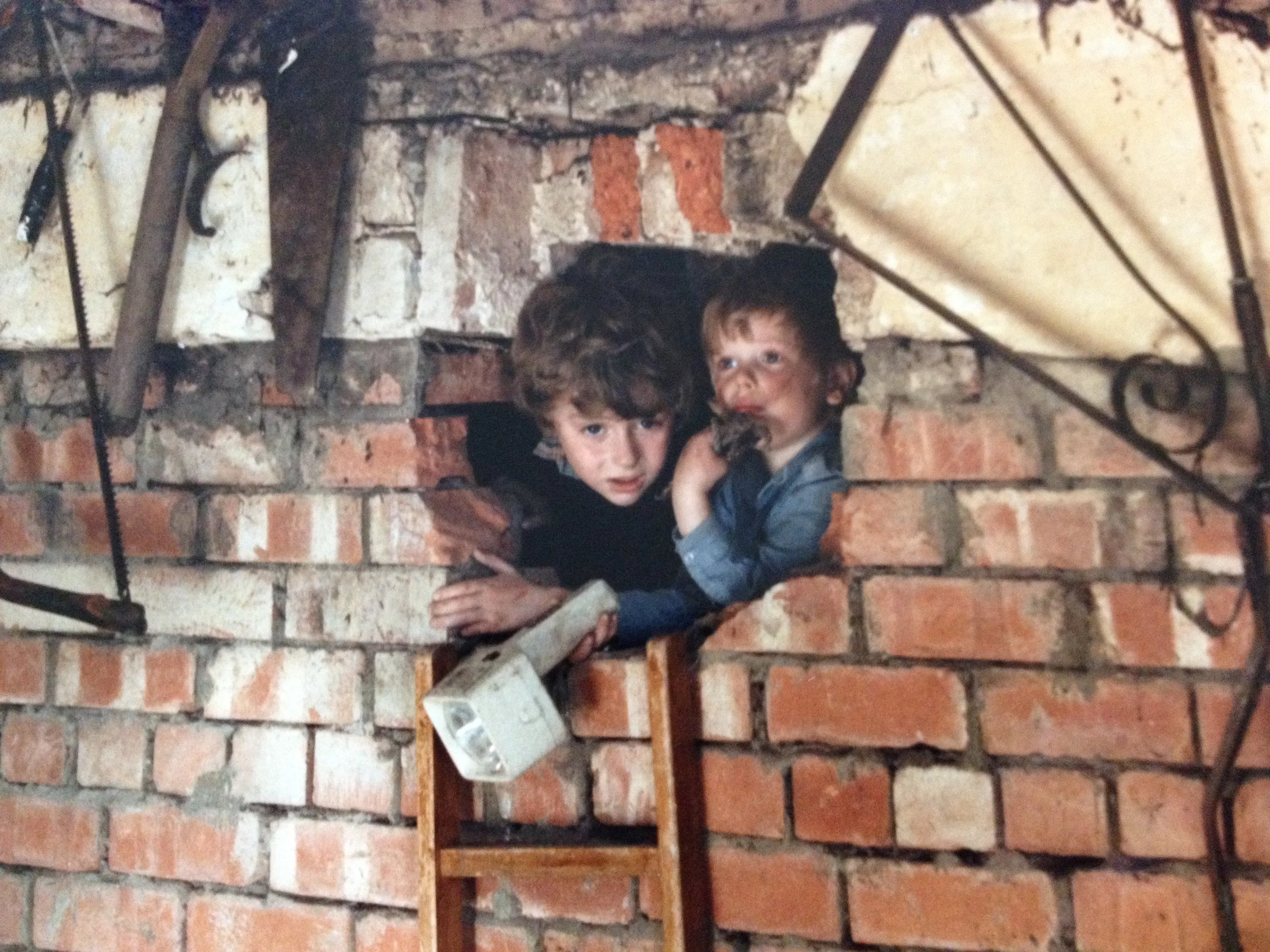Two young boys peek out from a small brick opening in a brick wall, surrounded by various tools and scaffolding, with a look of curiosity on their faces.