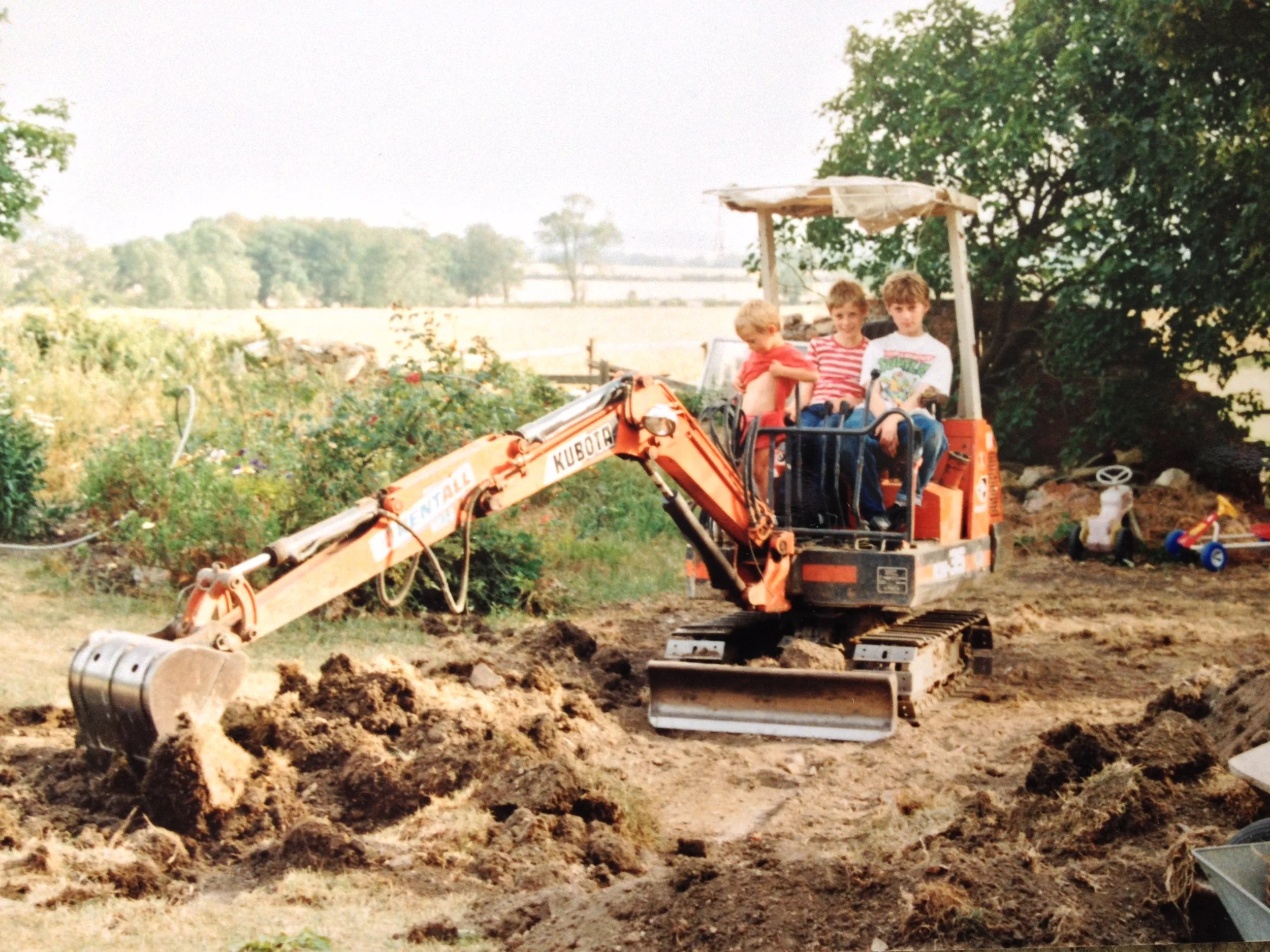 Three boys sitting on a toy excavator in a garden or yard, with trees and open land in the background, and toys scattered on the ground nearby.