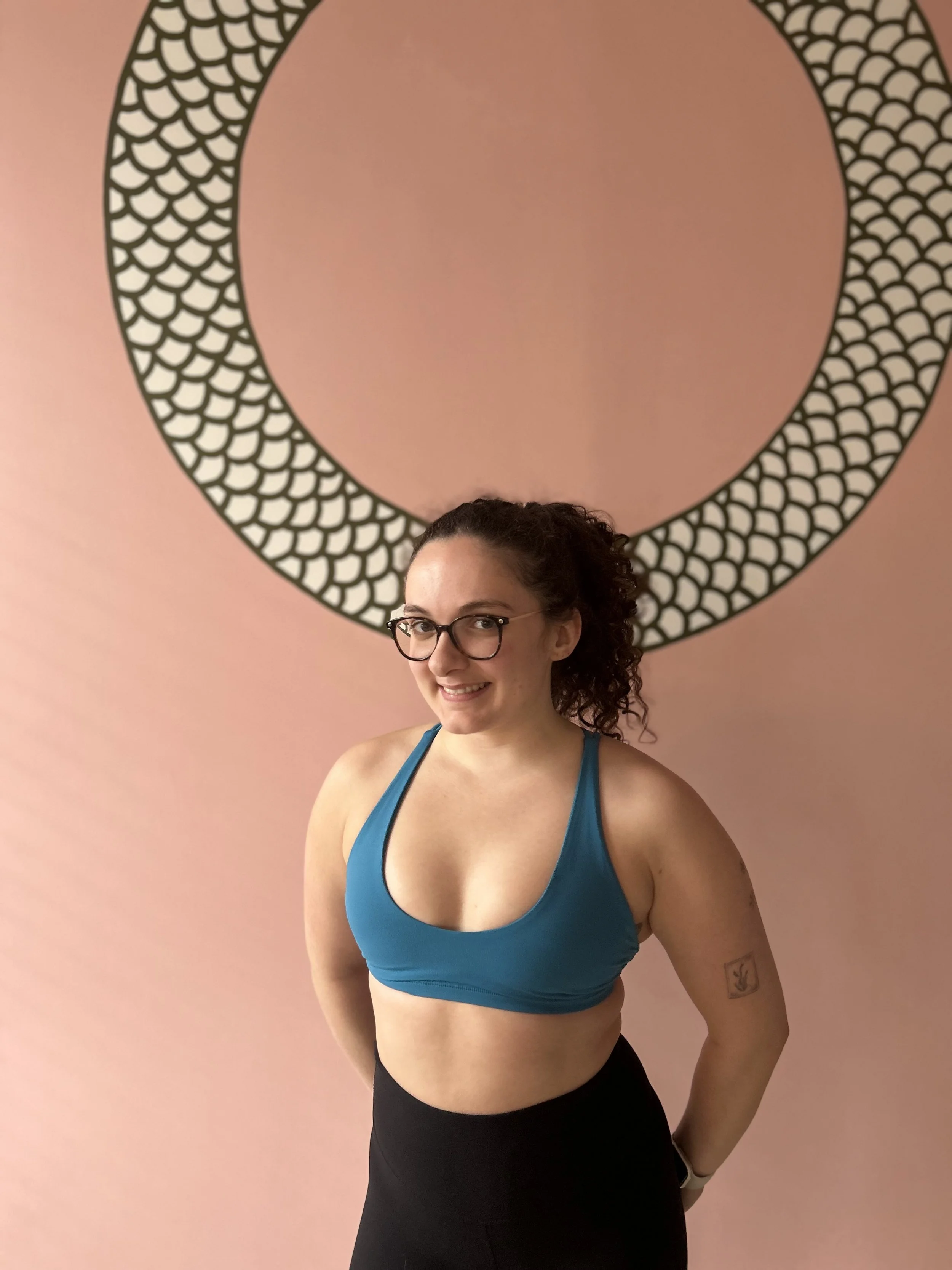 A person wearing a blue sports bra and black leggings stands in front of a pink wall with a circular pattern. They have glasses and curly hair tied back.