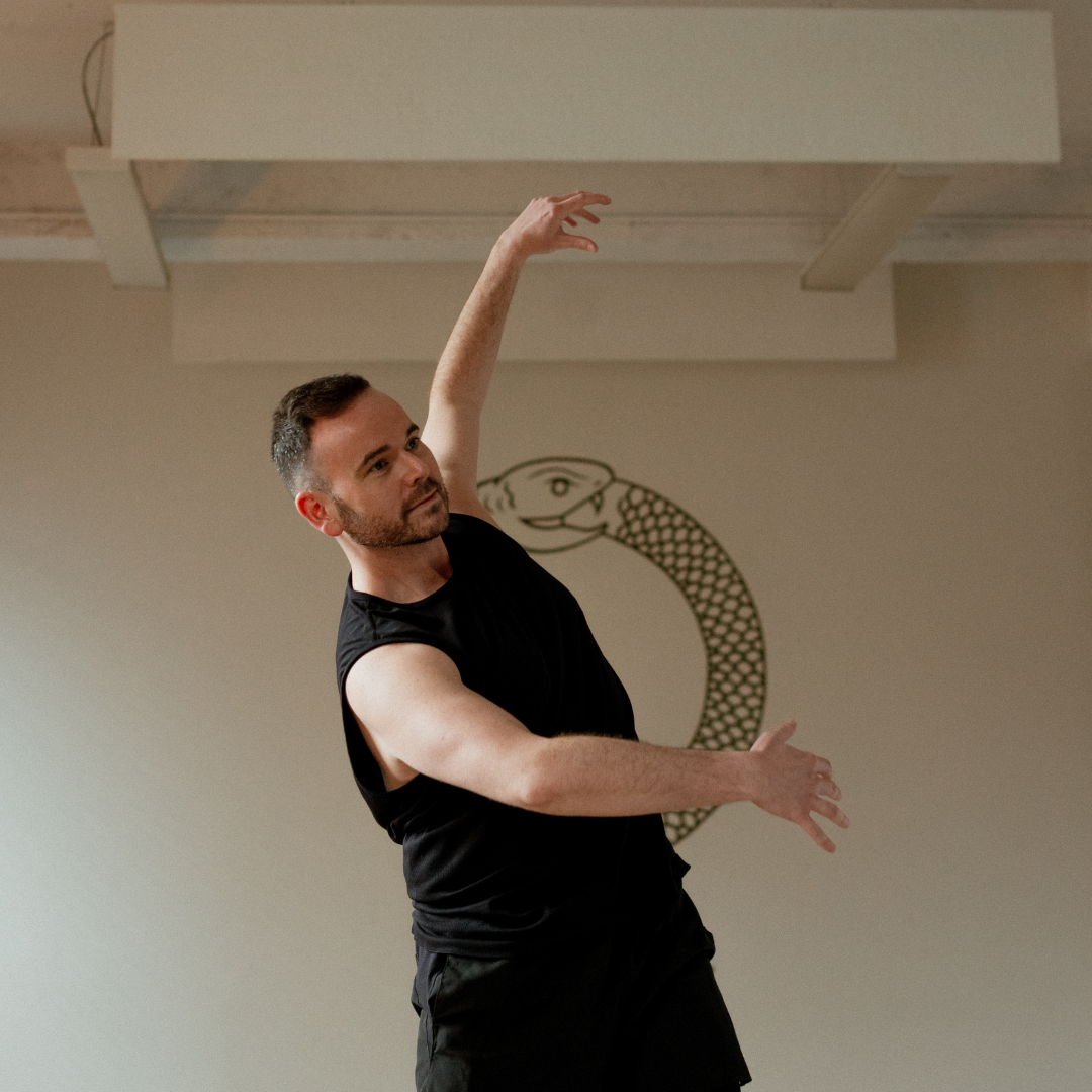 Person dancing indoors, wearing a black sleeveless shirt, posing with arms gracefully extended overhead.