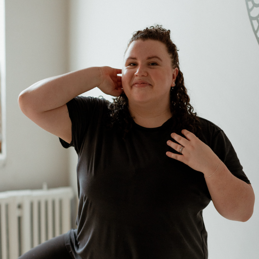 Person with curly hair smiling in a black t-shirt, one hand touching neck, standing indoors.