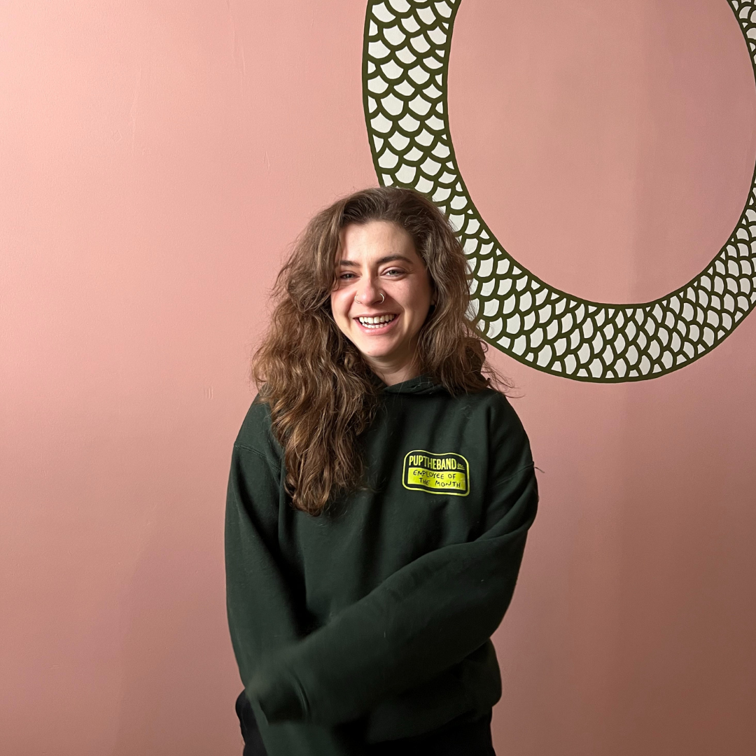 Person with long brown hair wearing a dark green hoodie stands smiling against a pink wall with a decorative circular pattern.