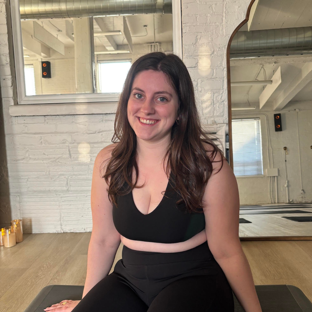 A woman sitting on a yoga mat in a fitness studio, wearing a black sports bra and leggings, smiling at the camera with a large mirror and white brick wall in the background.