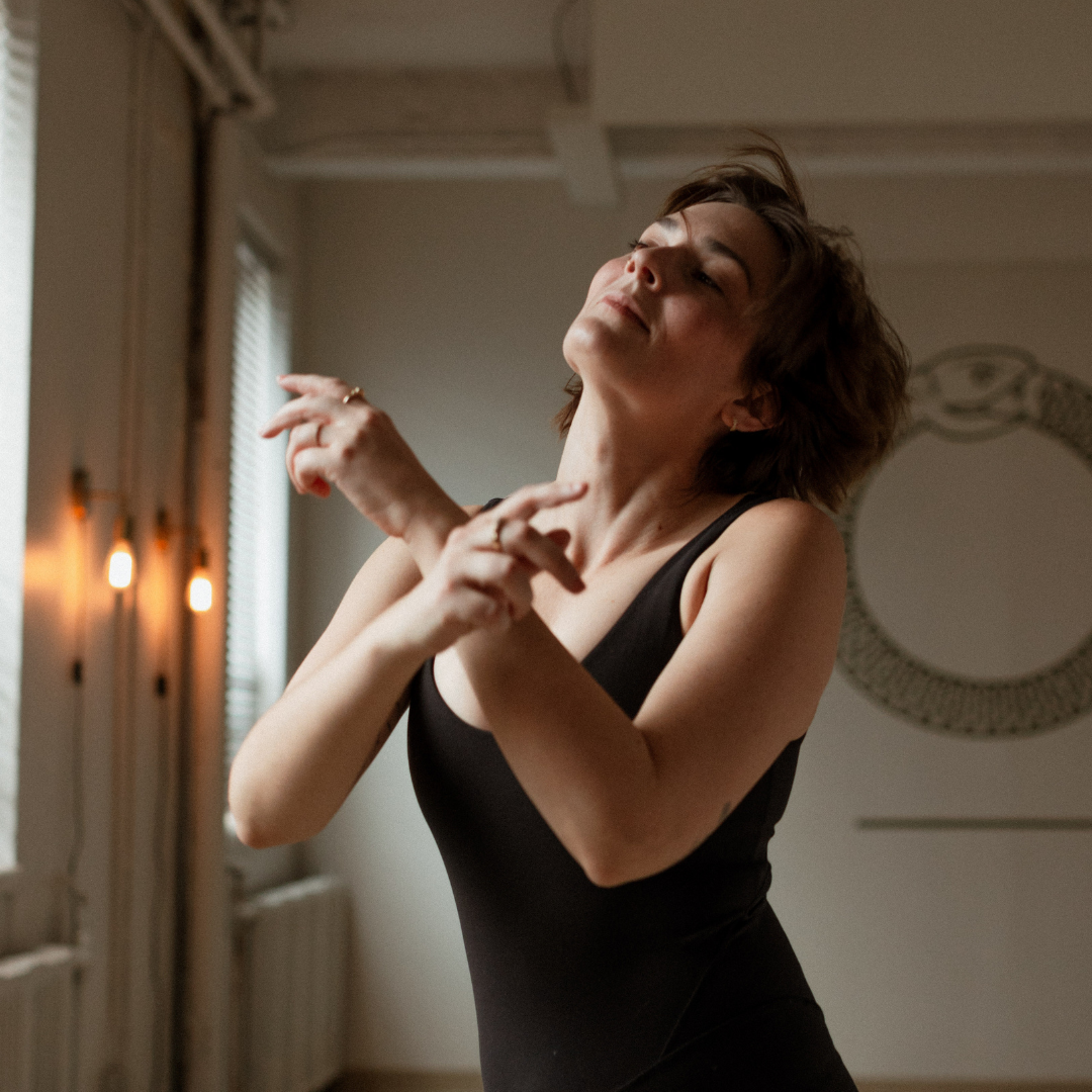 Woman dancing gracefully in a room with soft lighting and wall art in the background, wearing a black sleeveless top.