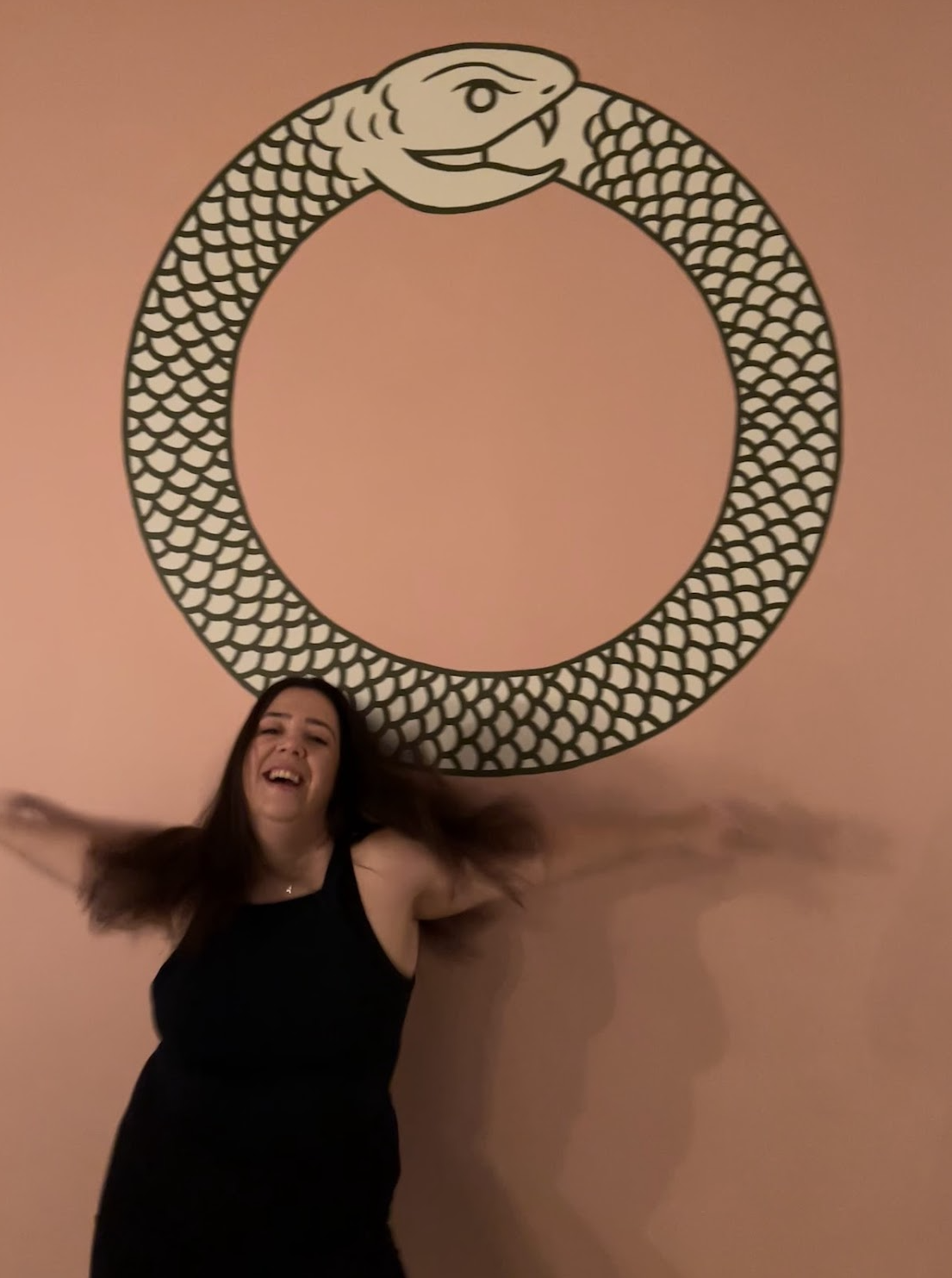 Person smiling with arms outstretched standing in front of a snake drawing forming a circle on a pink background.
