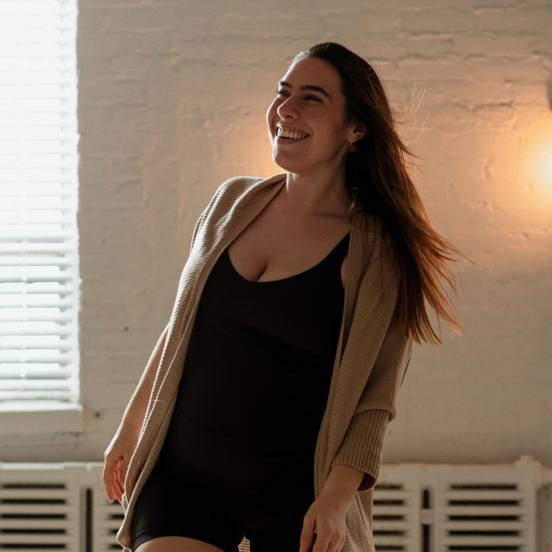 Woman smiling and looking away, wearing a black outfit and beige cardigan, standing in a room with a brick wall and soft lighting.