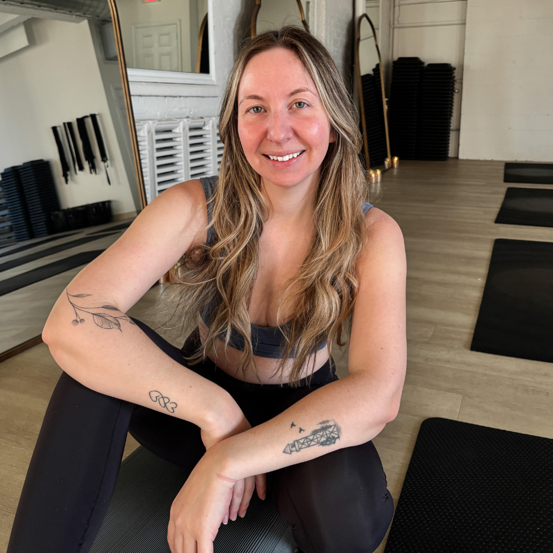 Smiling woman sitting on a yoga mat in a fitness studio with wooden floors, surrounded by exercise equipment. She has long hair and visible tattoos on her arms.