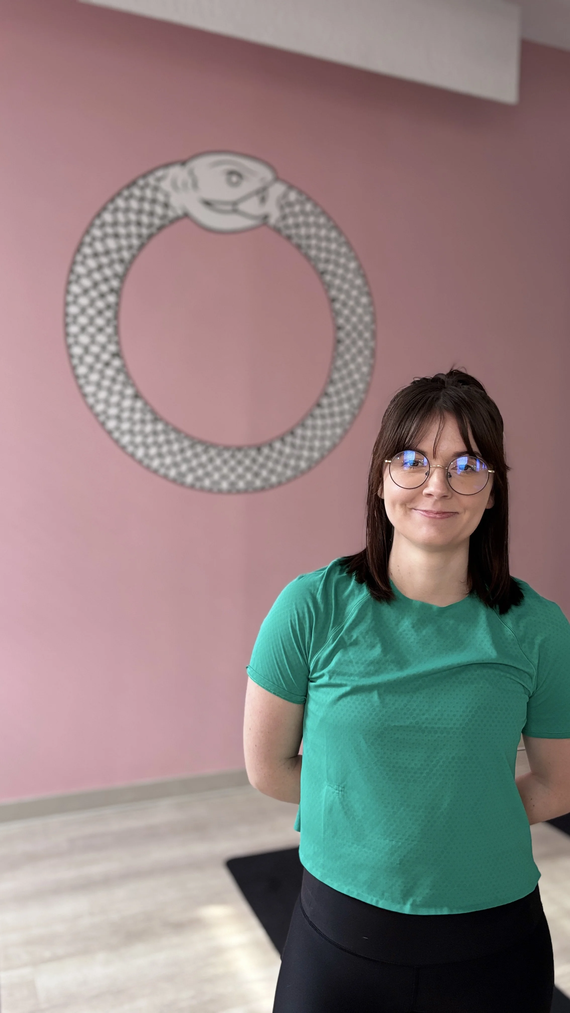 Person wearing glasses and a green shirt standing in front of a pink wall with a circular snake artwork.