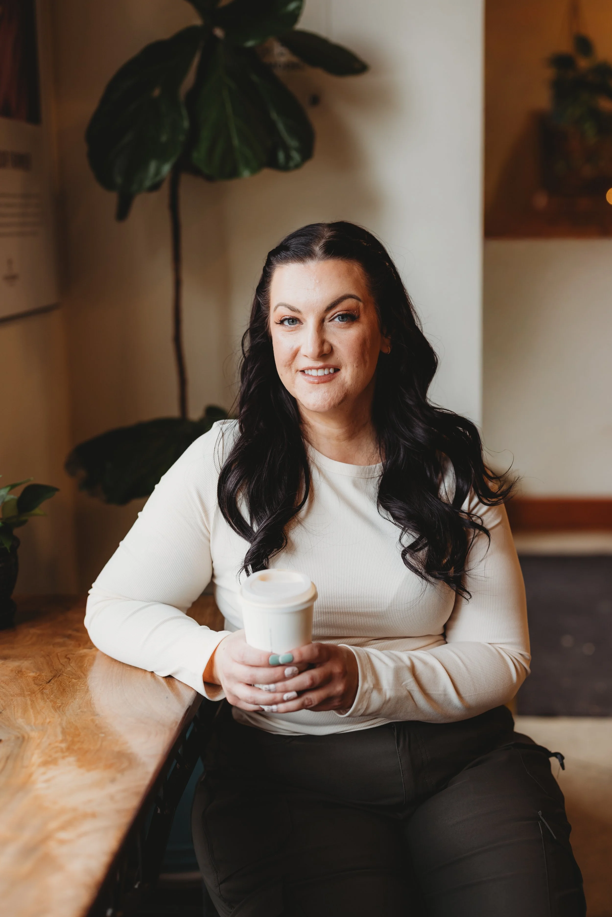 A woman with long dark wavy hair sitting at a wooden table, holding a white takeaway coffee cup, smiling at the camera.