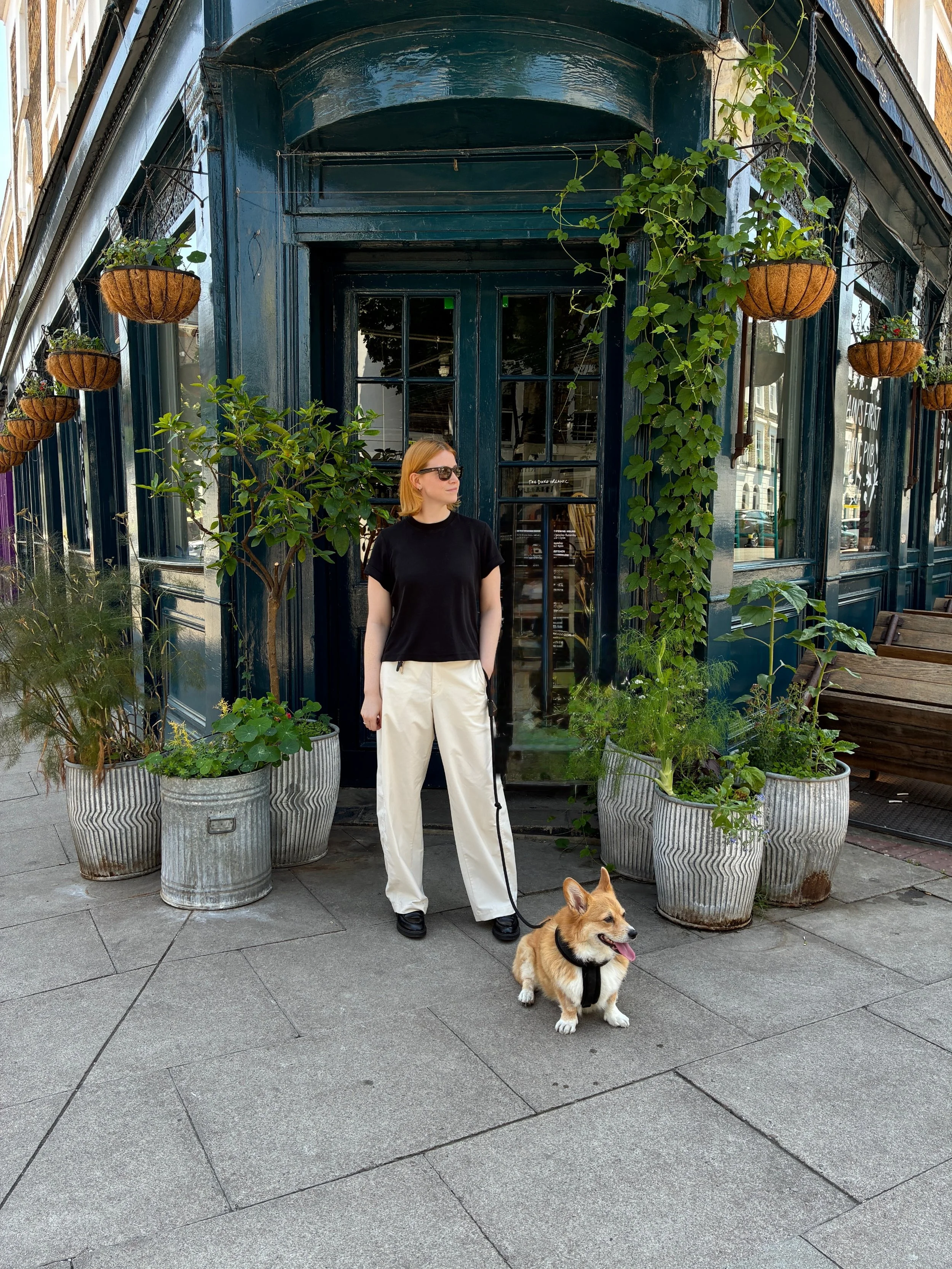 A woman with red hair wearing a black T-shirt, cream-colored pants, and black shoes standing outside a dark blue storefront with large windows. She is holding a leash attached to a small, tan and white dog sitting on the sidewalk. The storefront has hanging planters with plants, and large pots with green foliage surround the woman and dog.