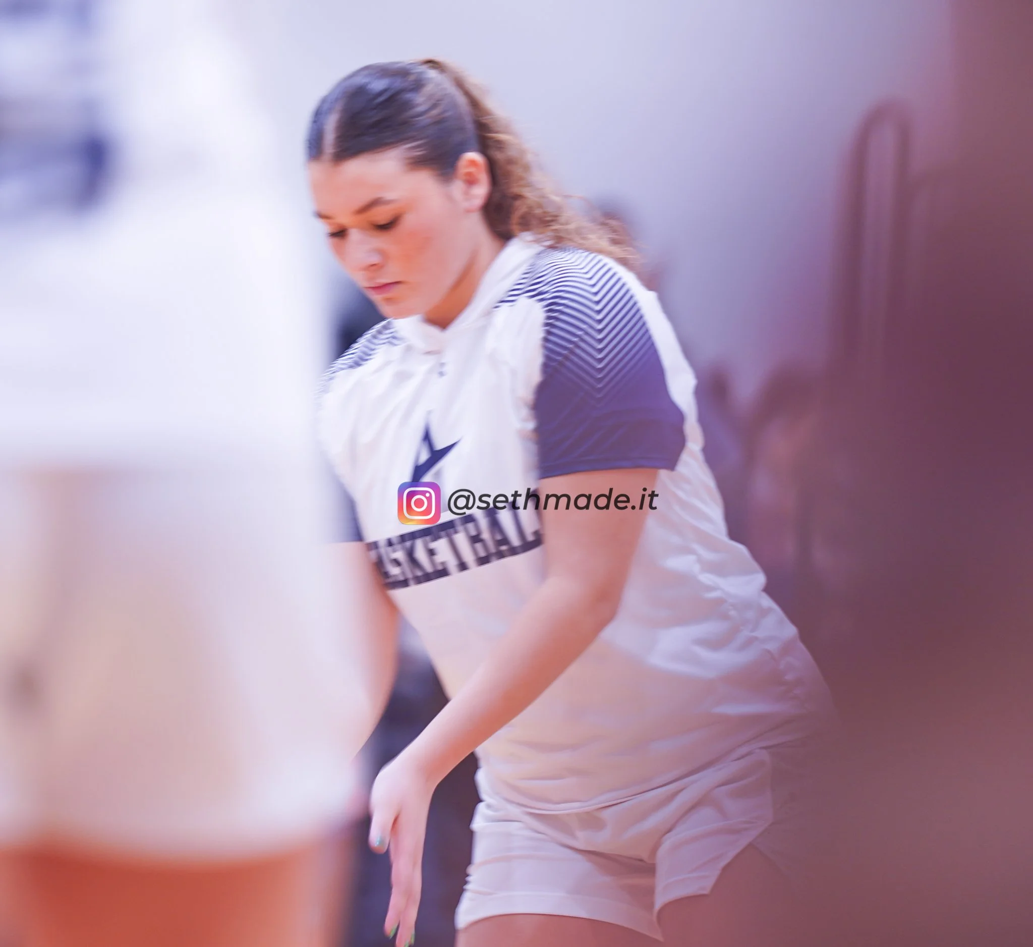 A female basketball player wearing a jersey with short black hair, looking down, in an indoor setting.