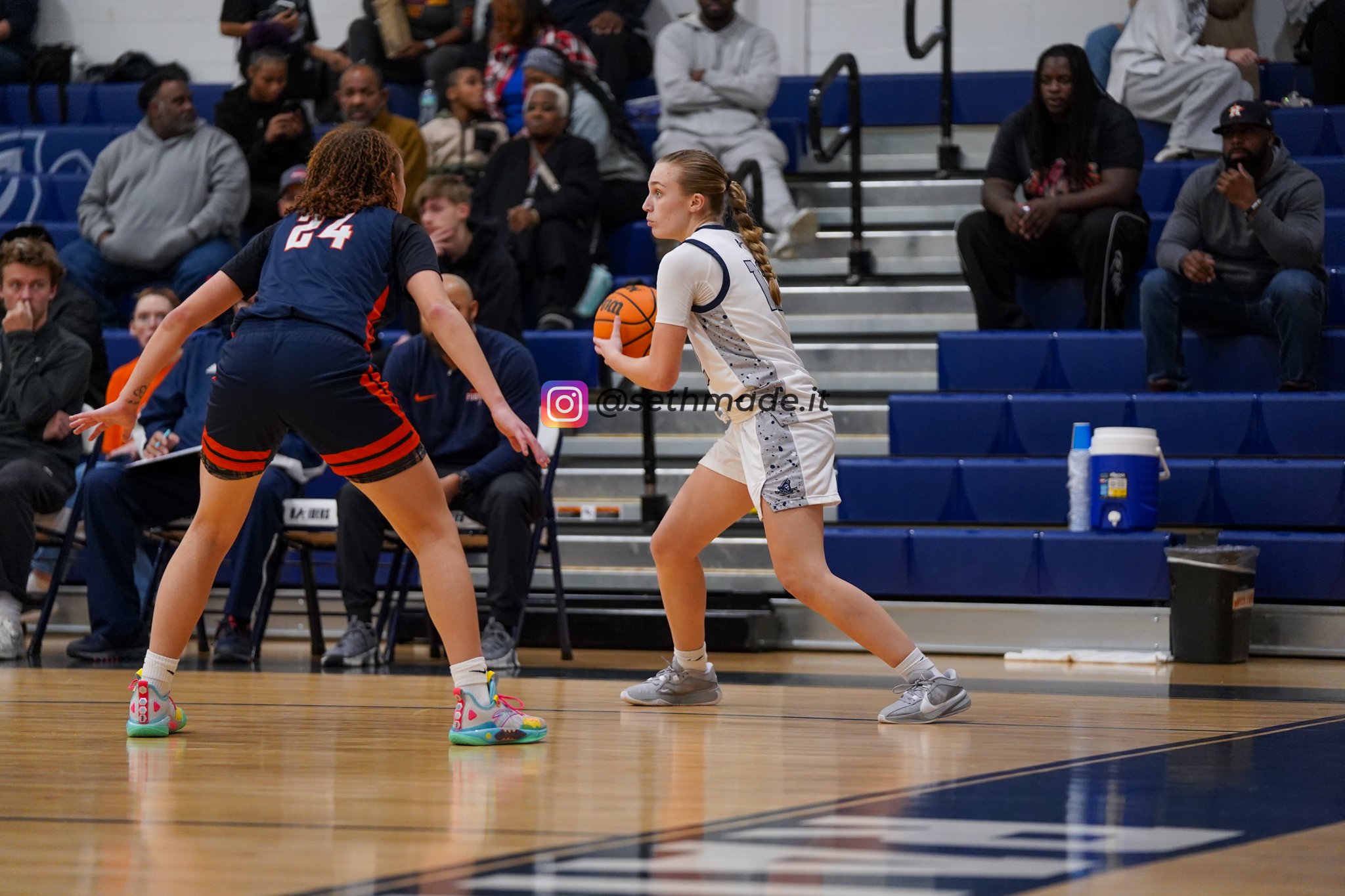A basketball game with one player holding a basketball and another player defending on the court, with spectators watching from the bleachers in the background.