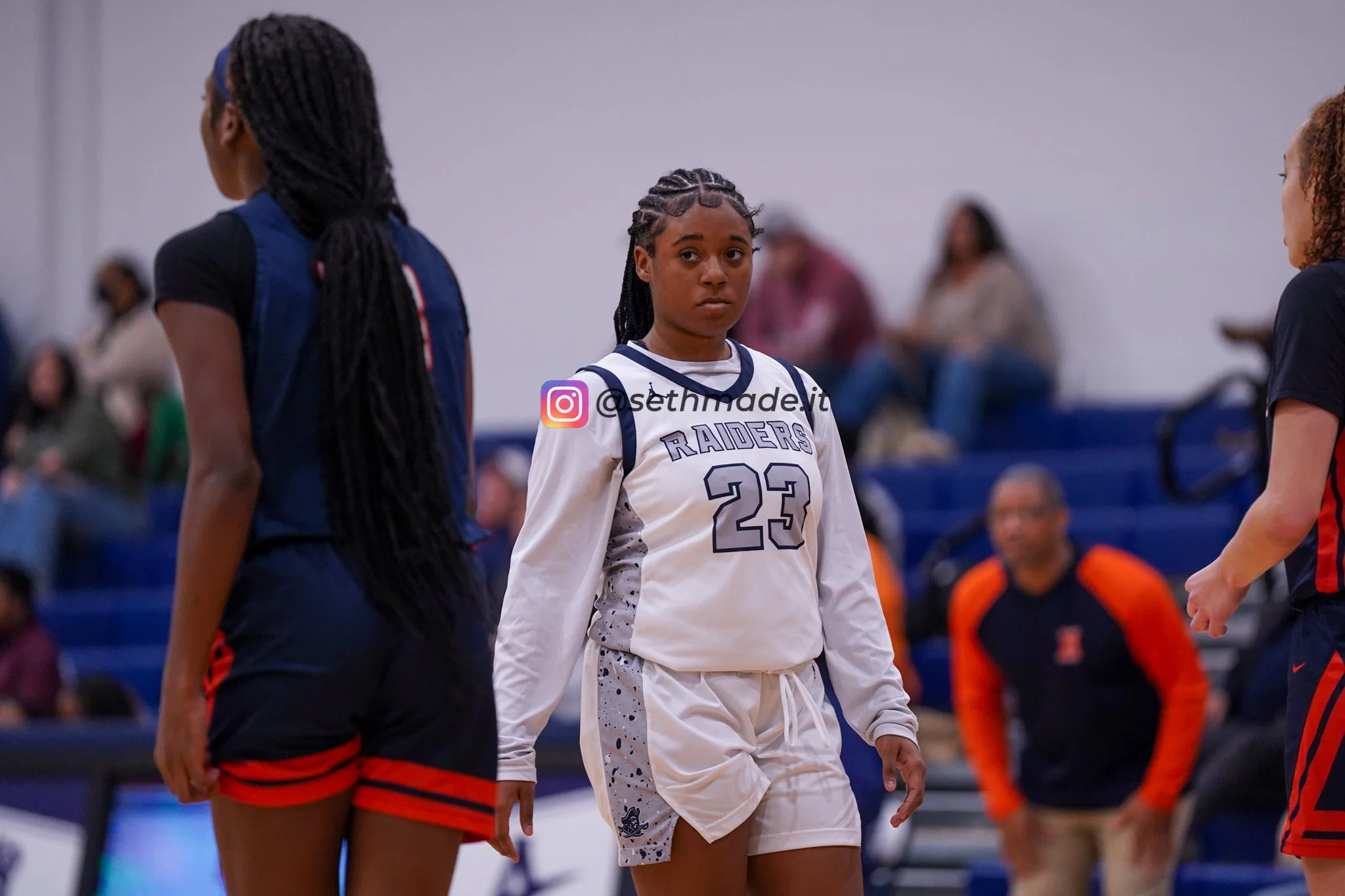 A young female basketball player in a white uniform with the number 23 on it, standing on a basketball court, looking serious. She is surrounded by other players and spectators in the background.