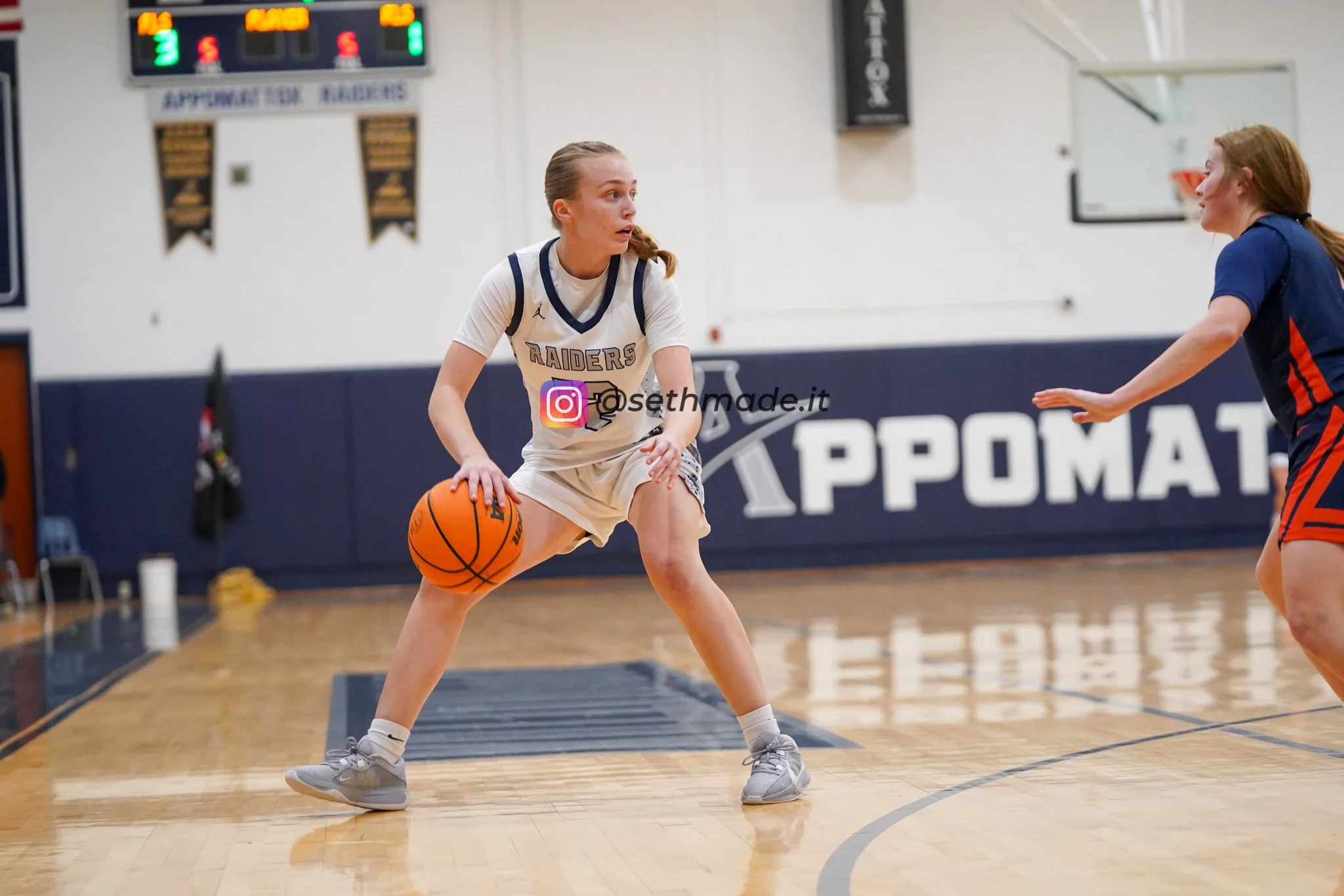 Two female basketball players on an indoor court, one wearing a white jersey with 'Raiders' and number 5, holding a basketball and guarding the other player, who is reaching out with her hand. The background features a scoreboard and banners.