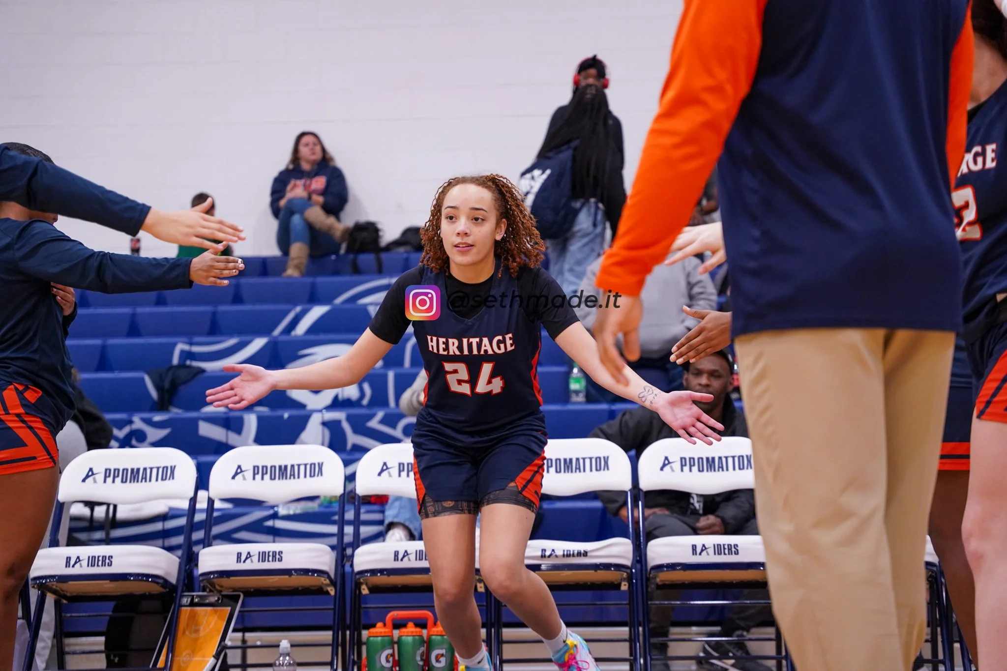 A young female basketball player with curly hair, wearing a black and red jersey with the number 24, is standing on a basketball court, extending her arms outward while other team members and coaching staff reach out to high-five or pat her on the ba