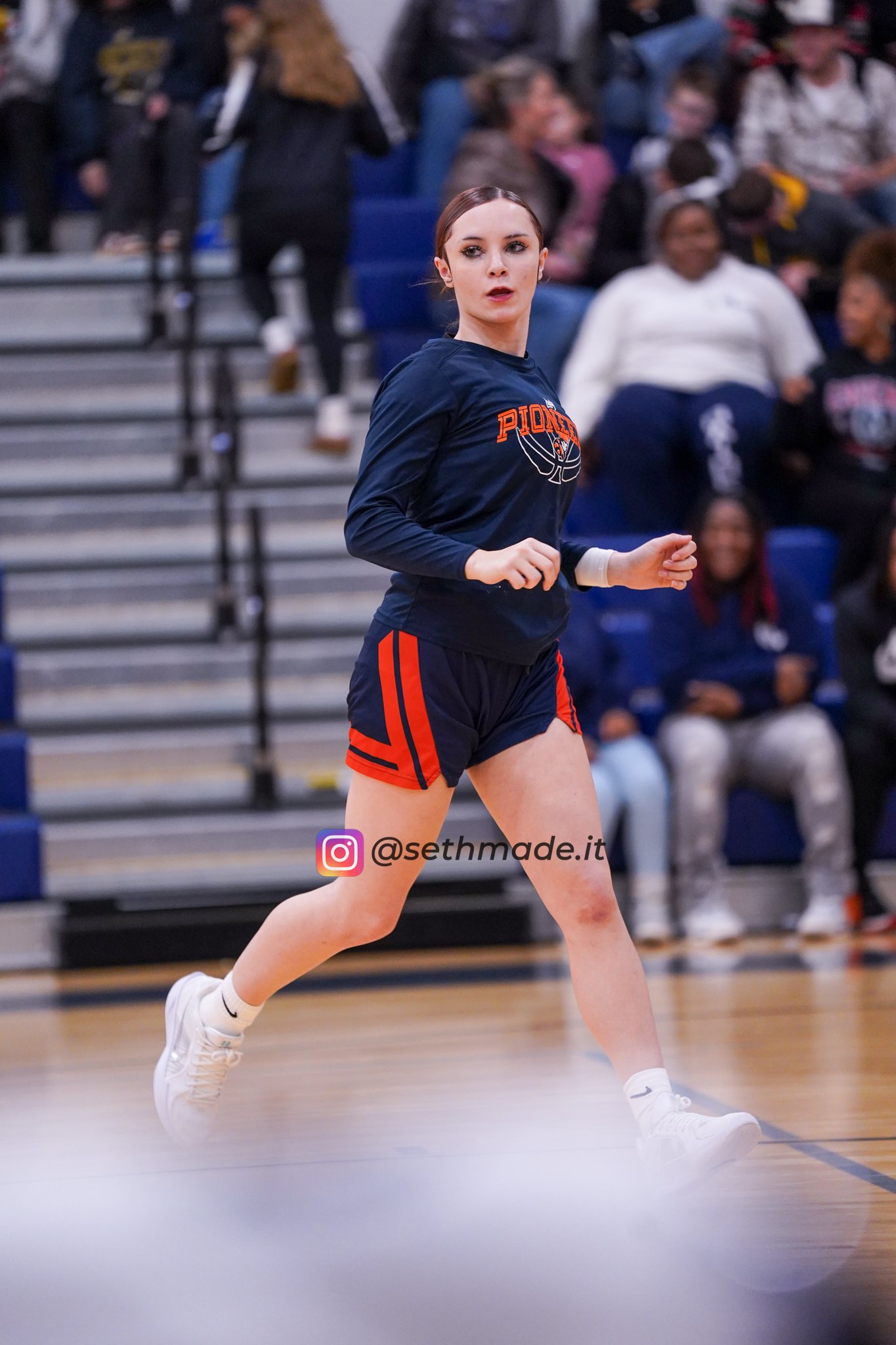 A young woman in athletic wear, with dark hair tied back, jogging on a gymnasium court during a sports event, with spectators seated on bleachers in the background.