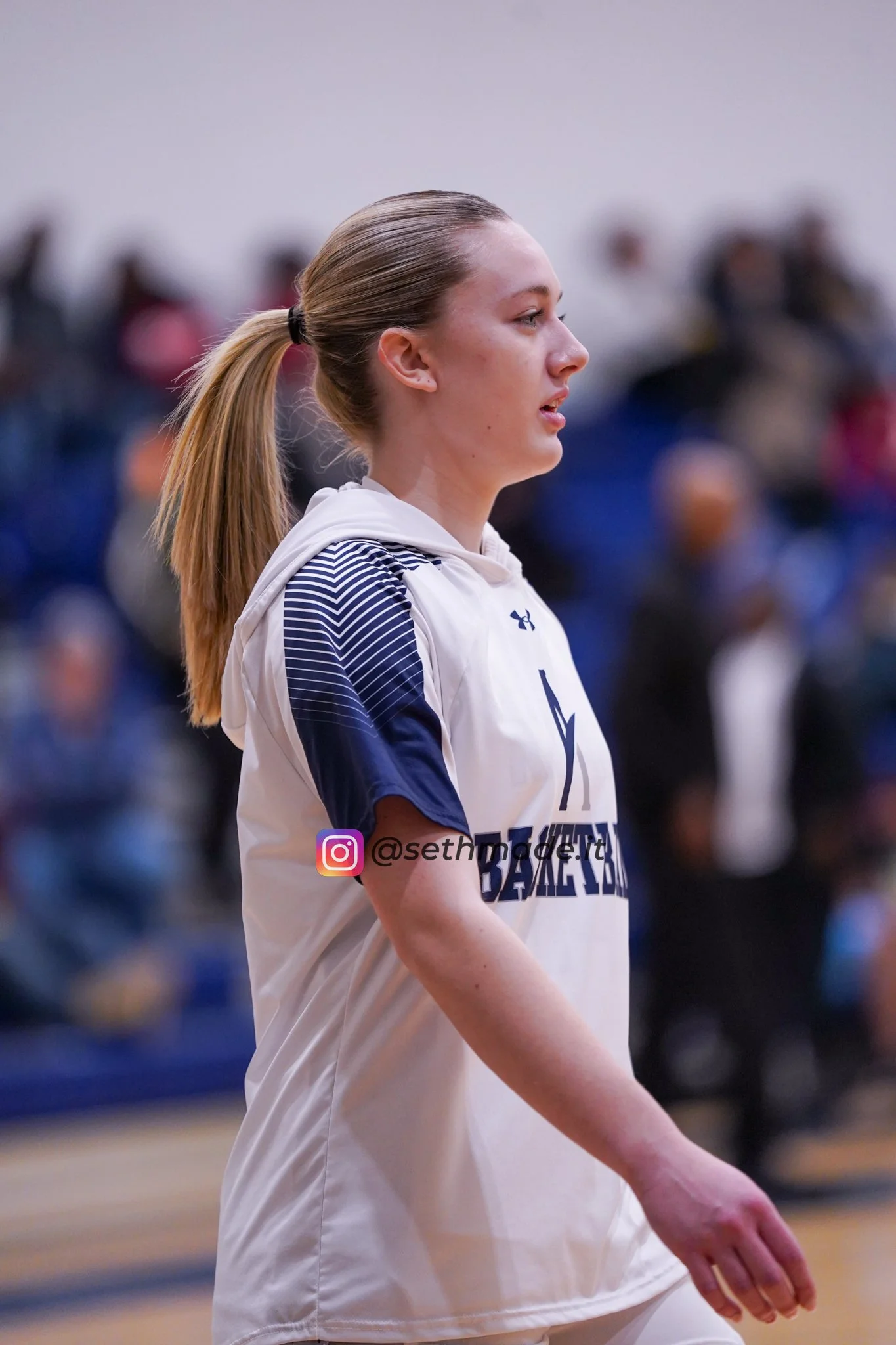Side profile of a young female athlete with a ponytail, wearing a white sports jersey with navy sleeves, standing on a basketball court.