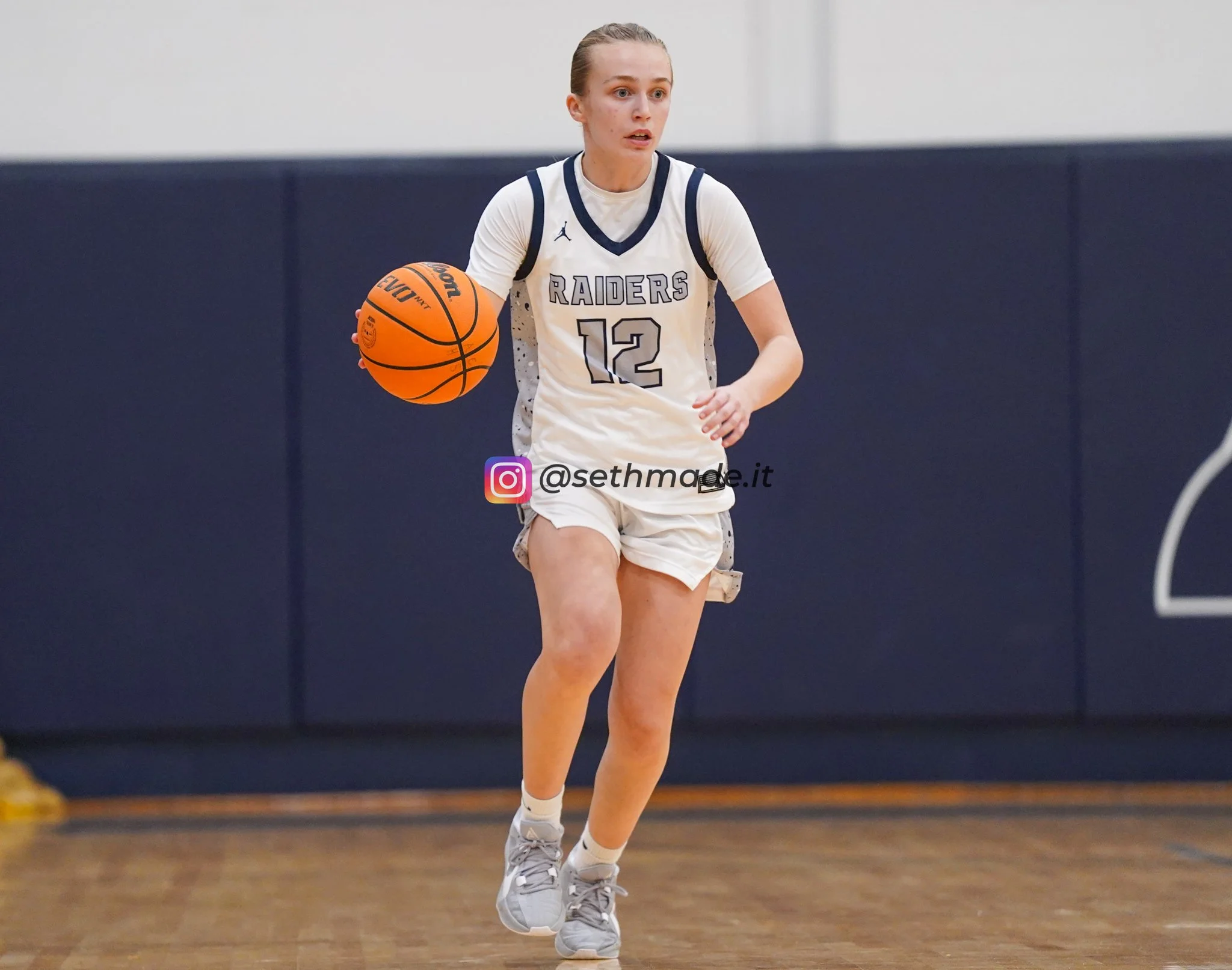 A female basketball player in a white jersey with blue accents and the number 12, dribbling a basketball on a gymnasium court.