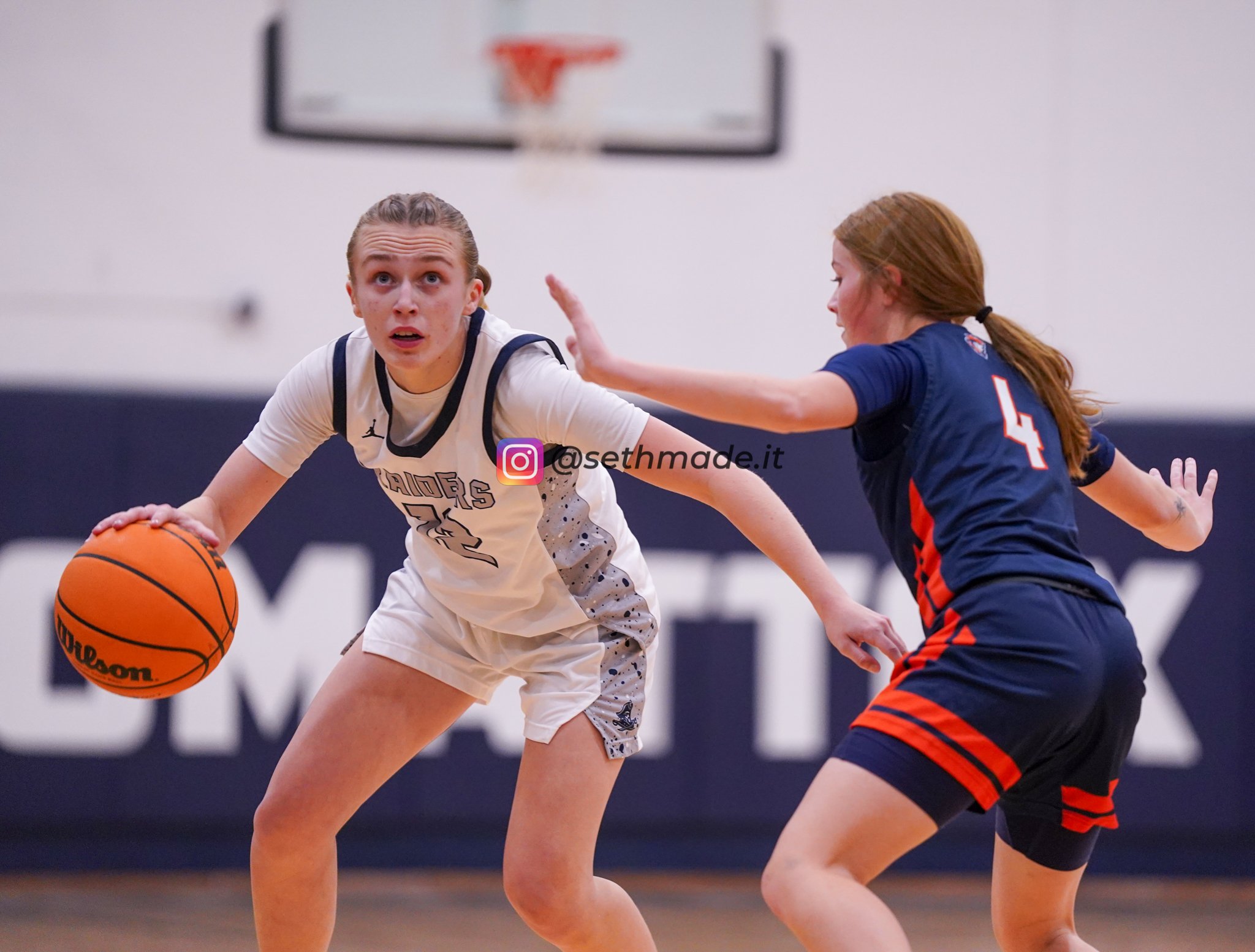 Two young female basketball players in action during a game, one in a white uniform dribbling the ball while the other in a navy blue uniform with red accents defends.