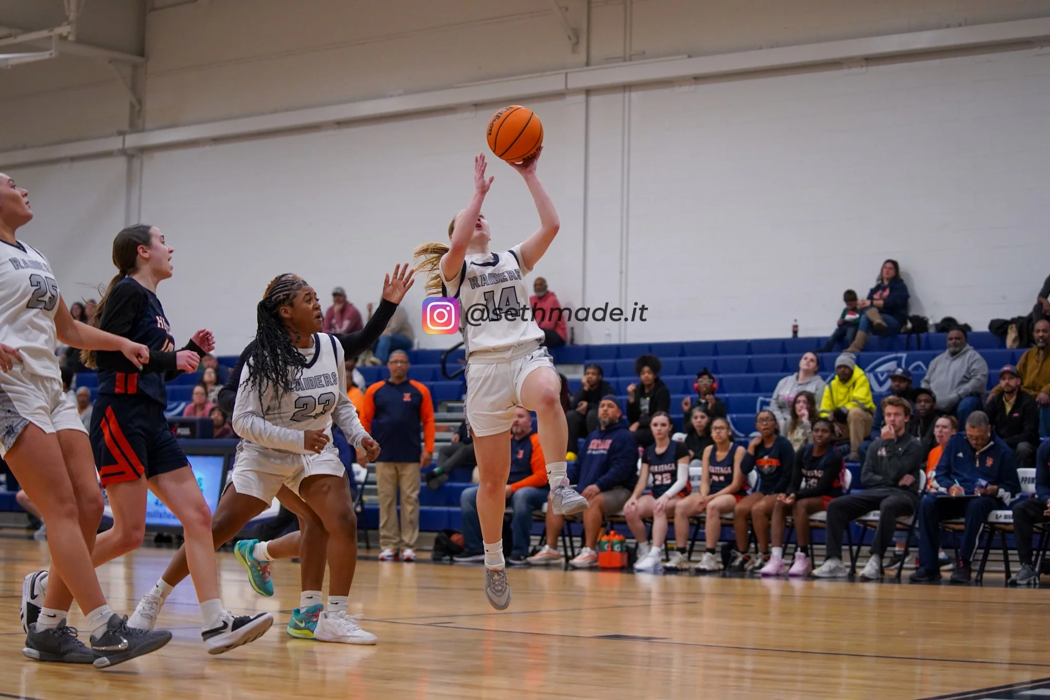A girl in a white basketball uniform with the number 14 jumps to make a shot during a game with opponents and spectators watching.