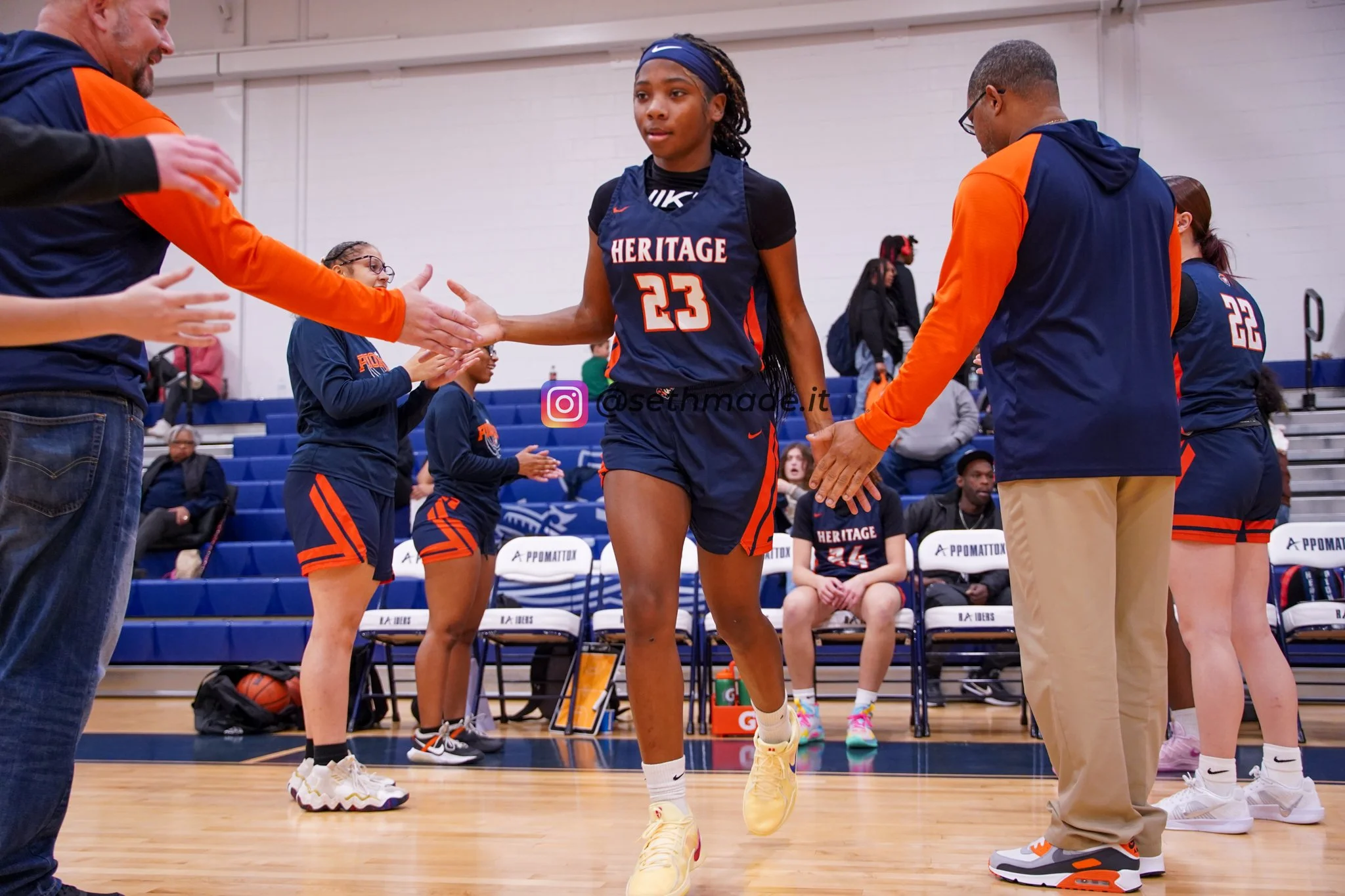 A young female basketball player performing a high-five with her coach during a game. She is wearing a navy basketball uniform with orange and white accents, and the number 23. Other team members and spectators are visible in the background.