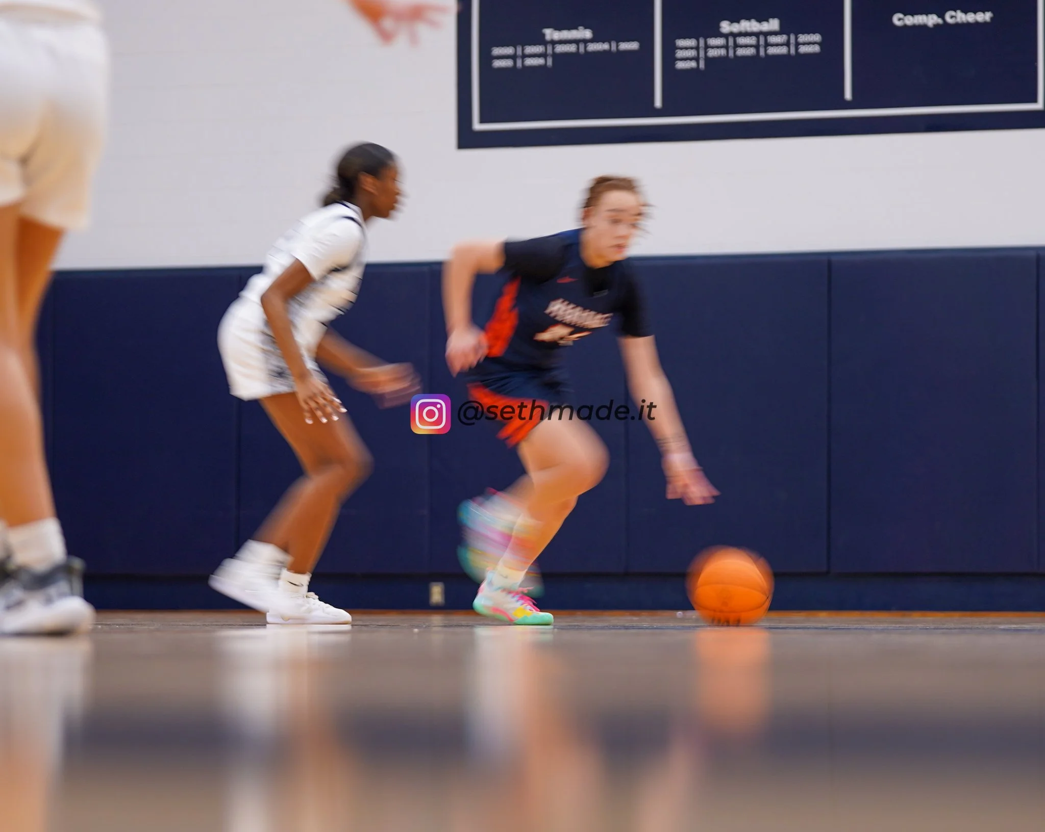 Two female basketball players in motion during a game in an indoor gym. One player in a white uniform and the other in a dark uniform is vying for control of a basketball near the ground. A blue wall with a scoreboard or poster is visible in the back