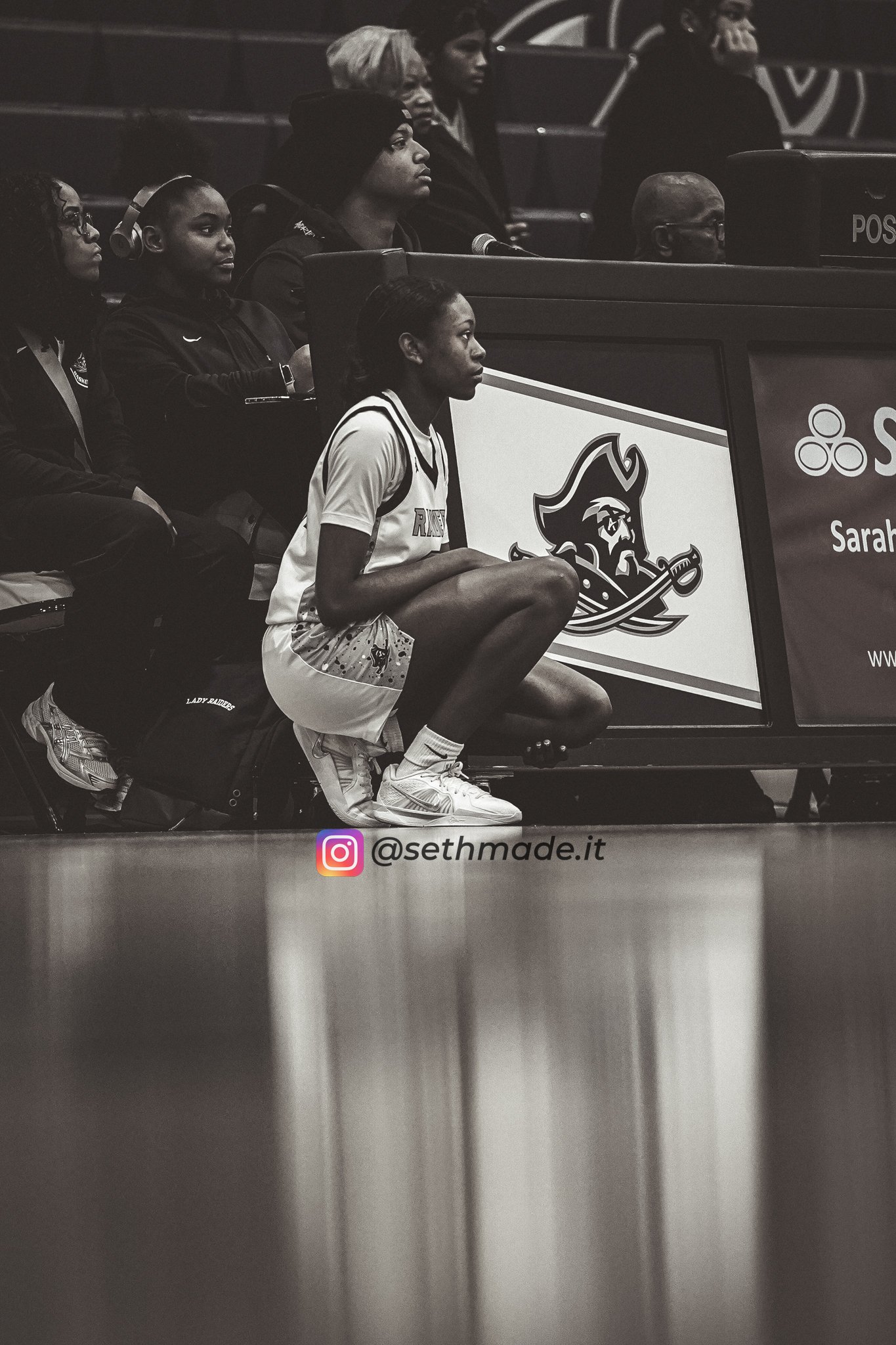 Black and white photo of female basketball players sitting on the sideline of a court. One player is crouched on the ground, dressed in a uniform and sneakers. Other players are sitting or standing nearby, with some wearing headphones. A large logo o