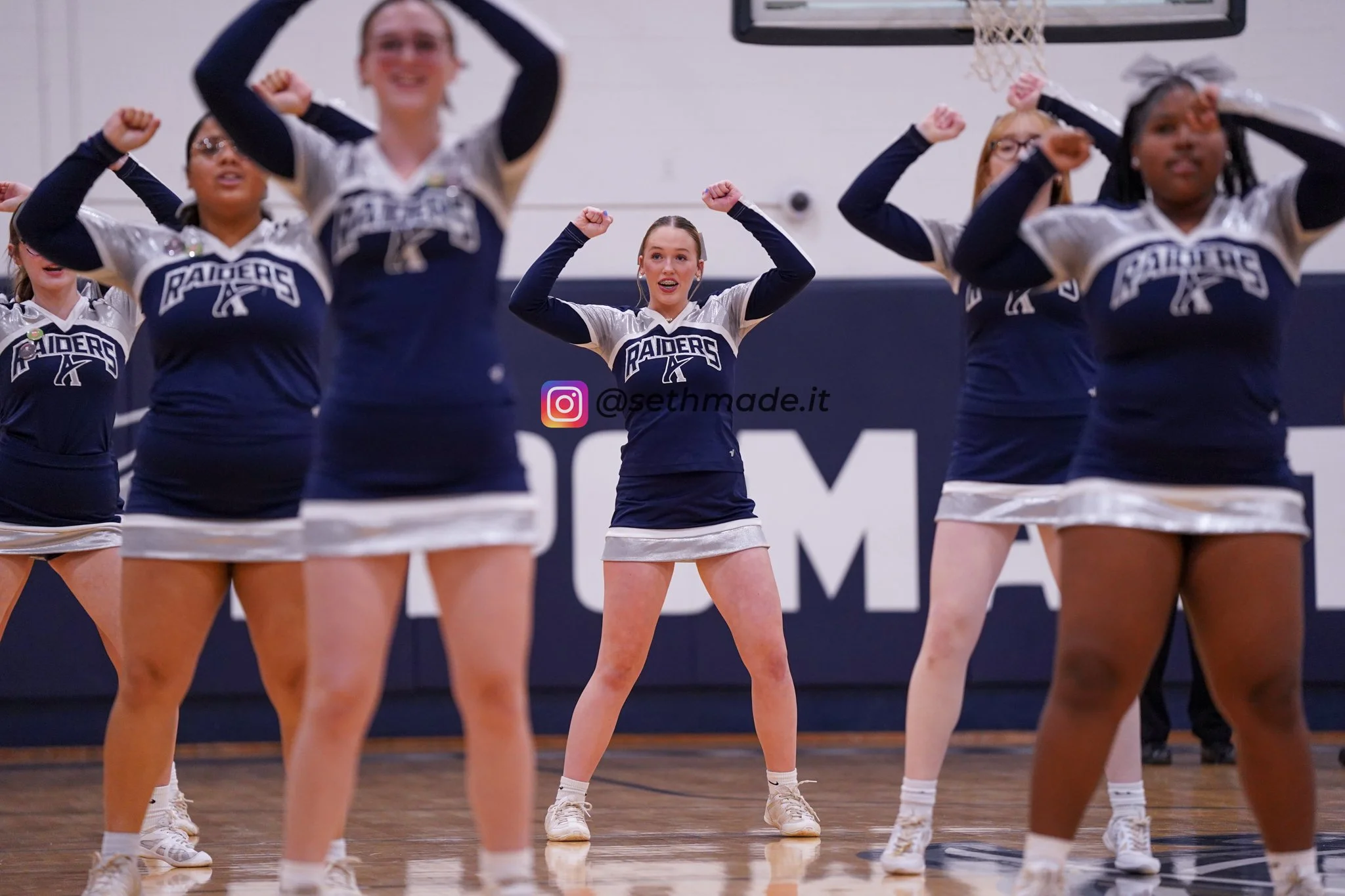 Cheerleaders in navy blue and silver uniforms performing a routine on a basketball court, with their arms raised and fists clenched.