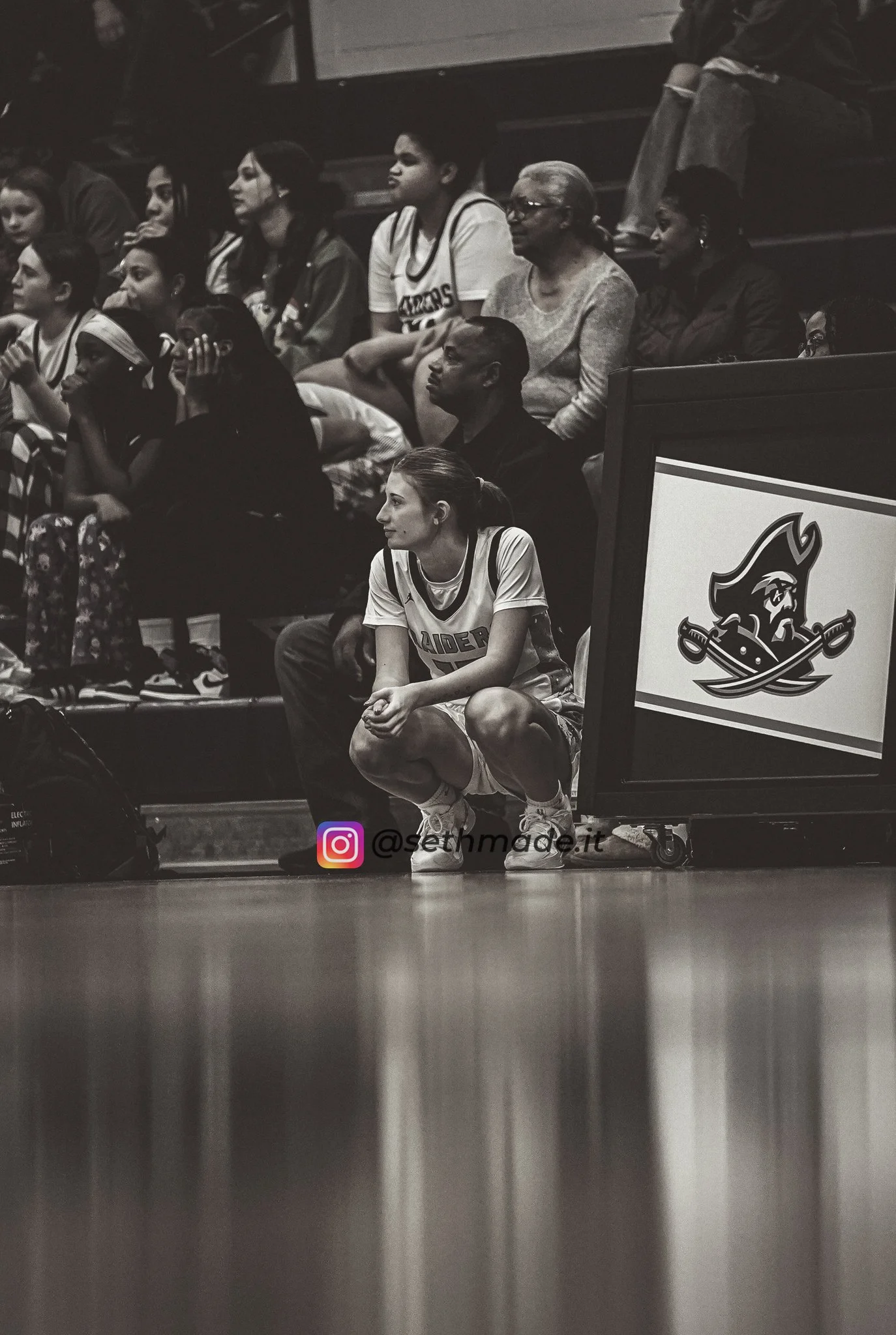 A female basketball player crouching on the sidelines during a game, with a group of spectators sitting behind her in the stands. The photo is in black and white and features a sign with a pirate logo on the court.
