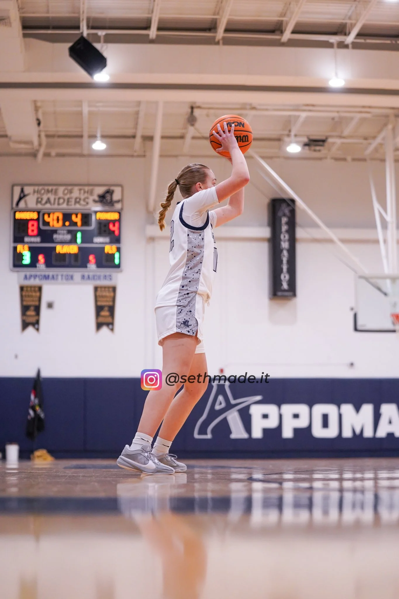 A female basketball player in a white uniform shooting a basketball in an indoor gymnasium.