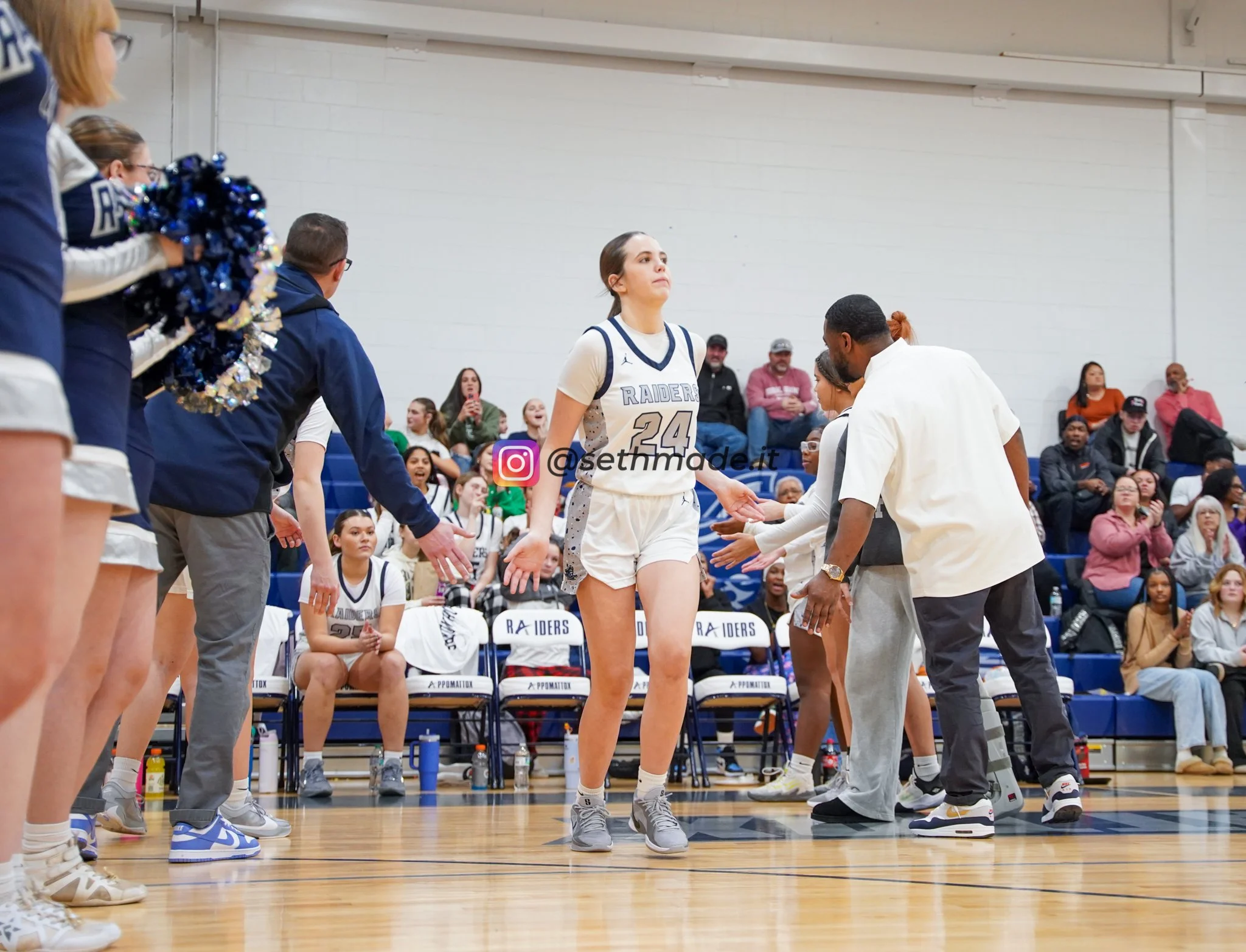 A basketball player wearing a white and gray uniform with the number 24 stands on the court, surrounded by teammates and coaches during a game or timeout, with spectators sitting in the bleachers in the background.