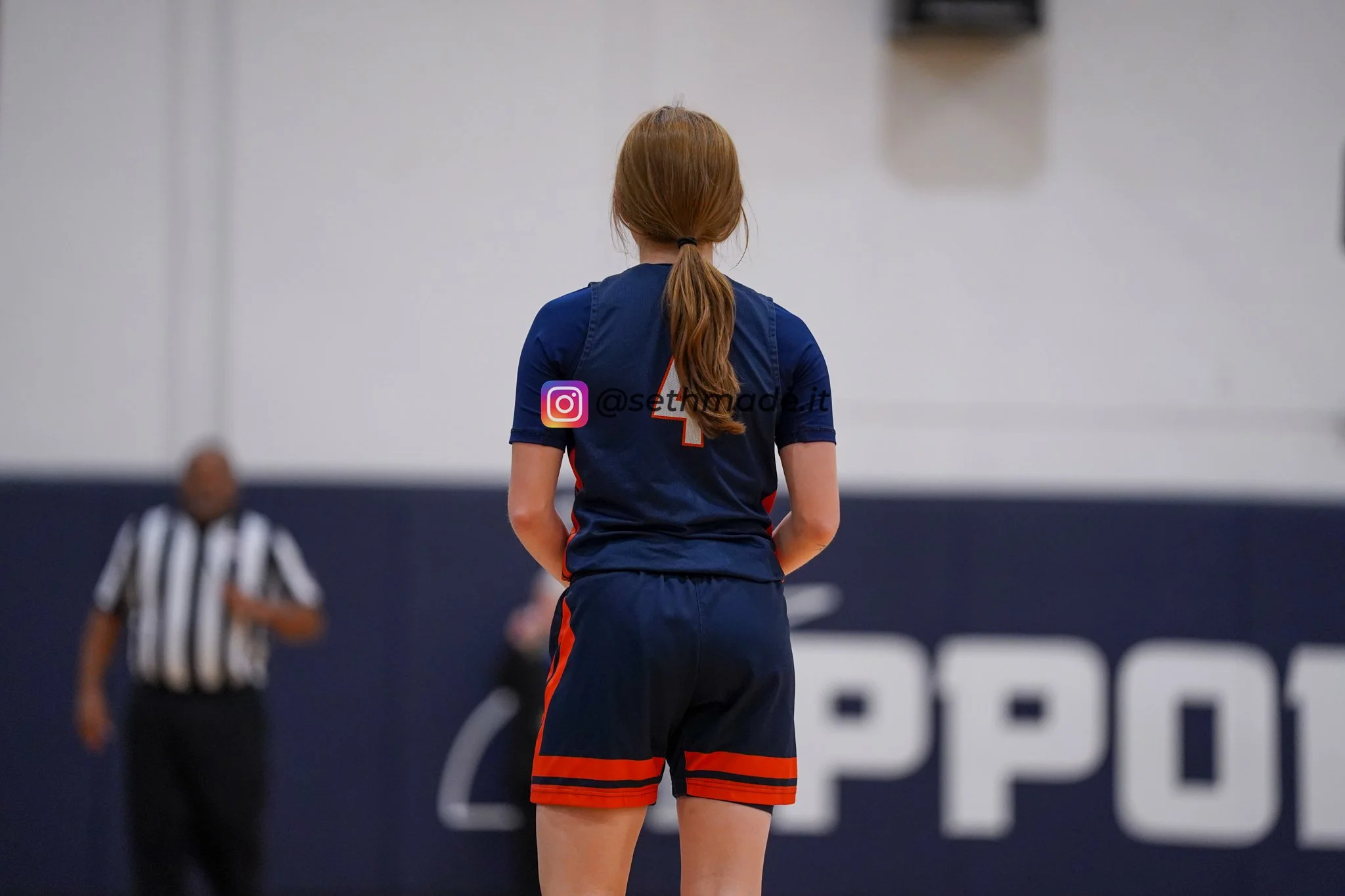 A female athlete with long red hair tied in a ponytail, wearing a dark blue sports uniform with a red number 4 on the back, standing on an indoor sports court with her back to the camera, facing a referee and other players.