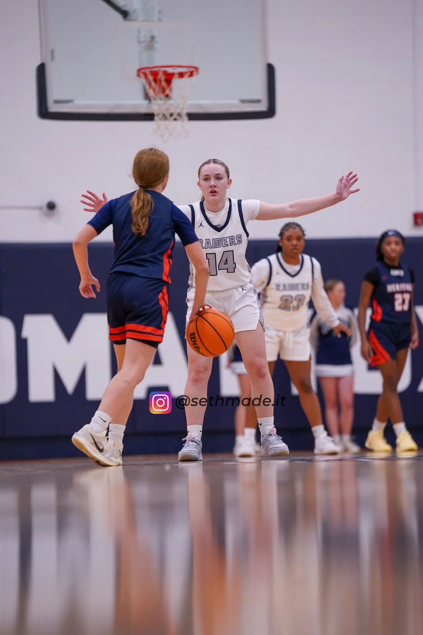 A basketball game featuring two young female players, one in a white uniform with number 14 and the other in a navy blue uniform with red accents, with other players and cheerleaders in the background.