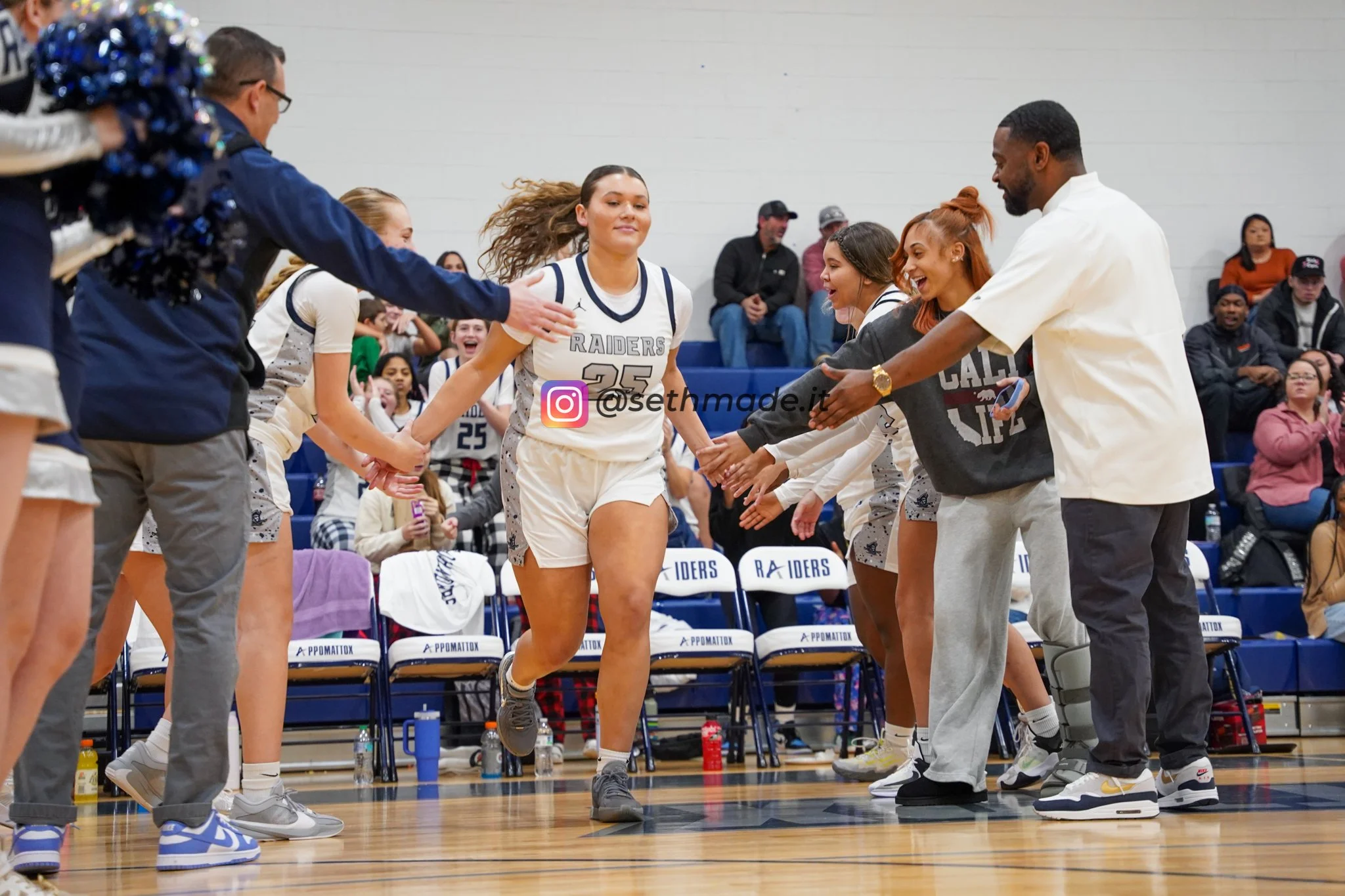 Women's basketball team lining up for a pregame or timeout introduction, with players shaking hands with coaches and staff on the court, and spectators watching in the background.