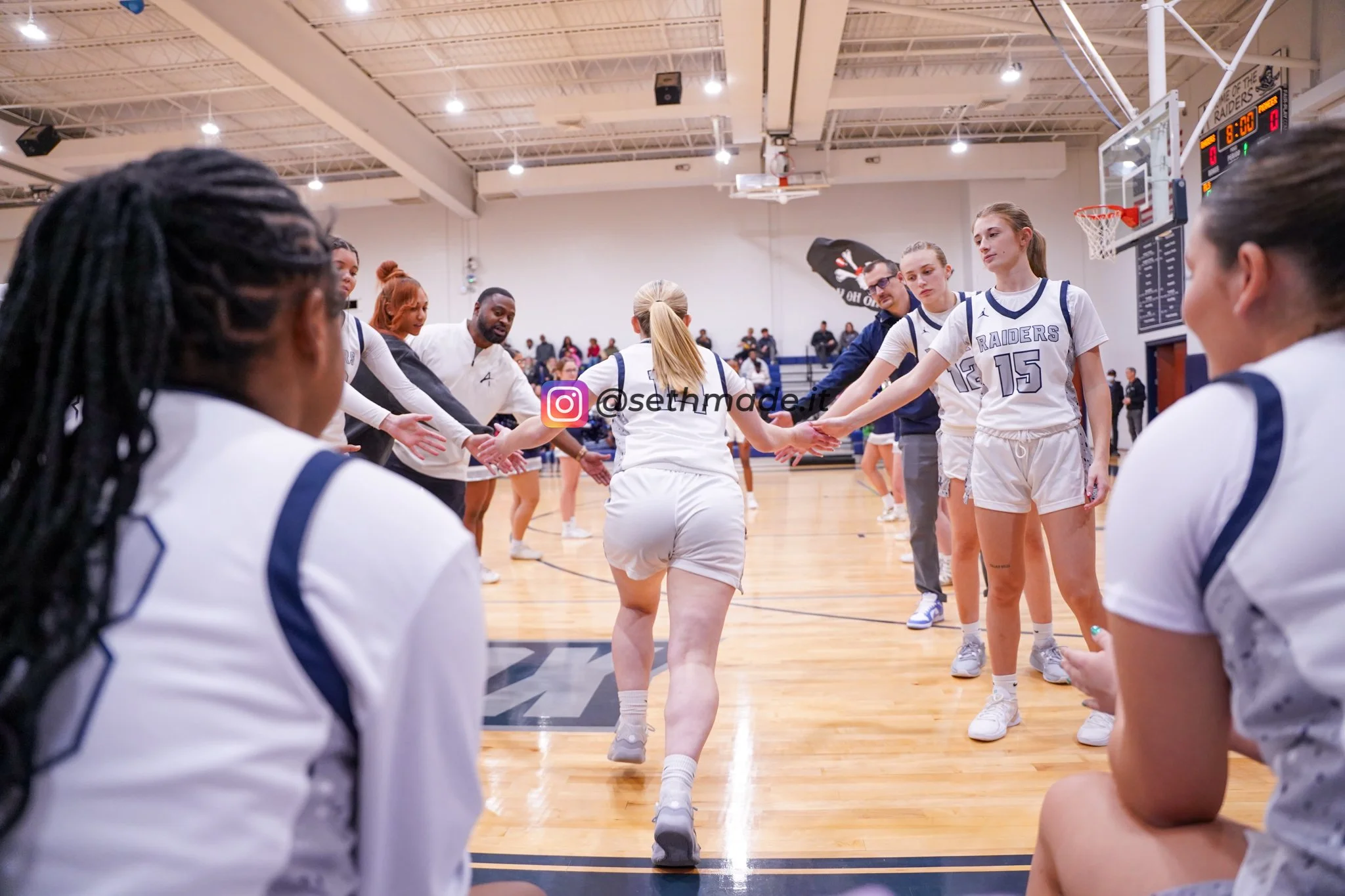 A women's basketball team standing in a circle on the court, holding hands before a game or practice in an indoor gymnasium, with spectators in the background.