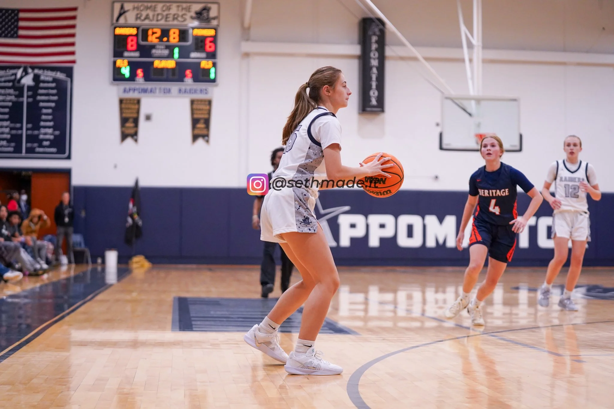 A female basketball player in a white jersey is preparing to shoot a free throw on a basketball court, with two other players and spectators in the background.